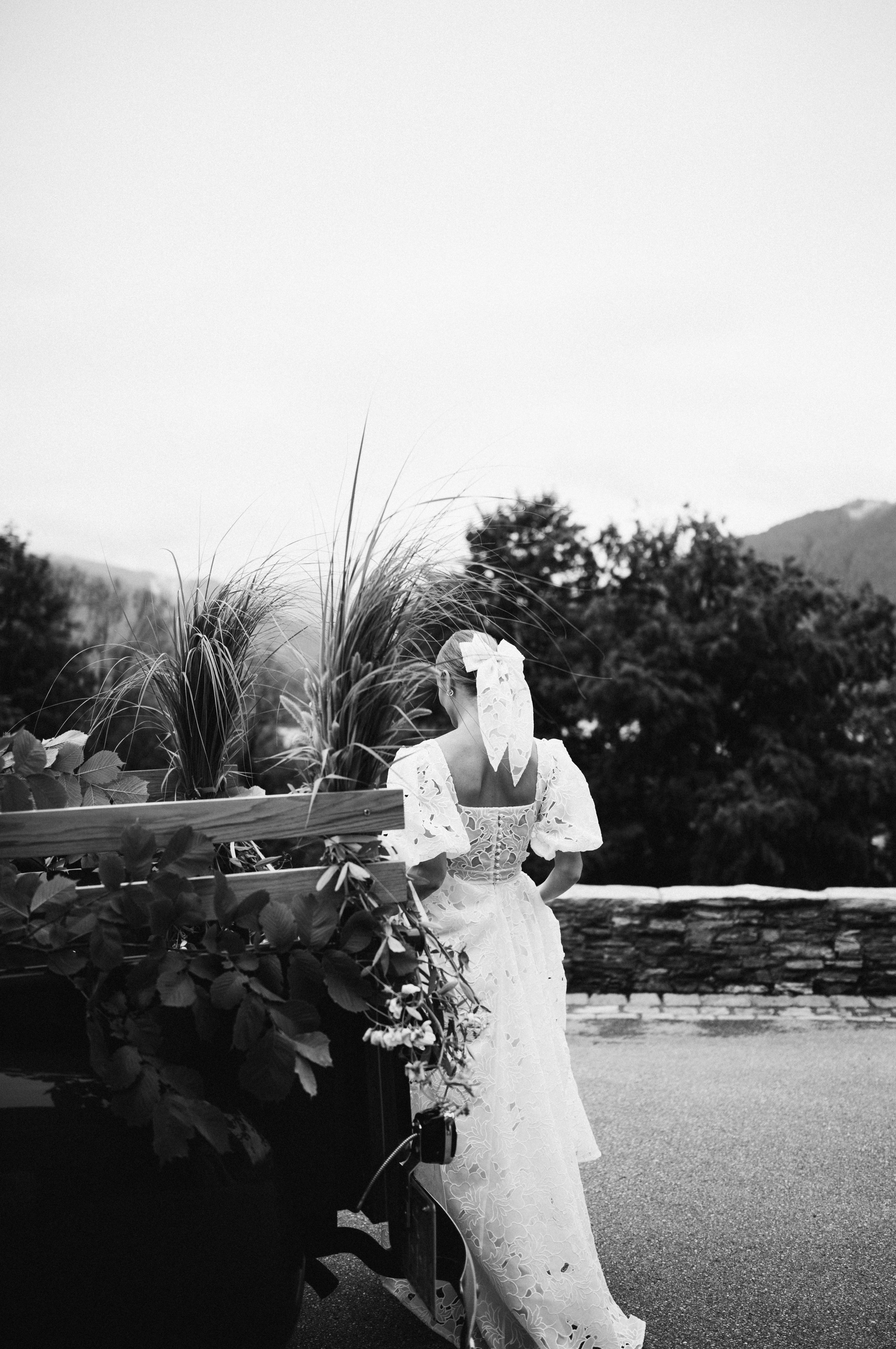 Black and white photo of a bride in a lace dress from behind, beside a vintage car with mountains beyond. Wedding photography by FADETOBLACK