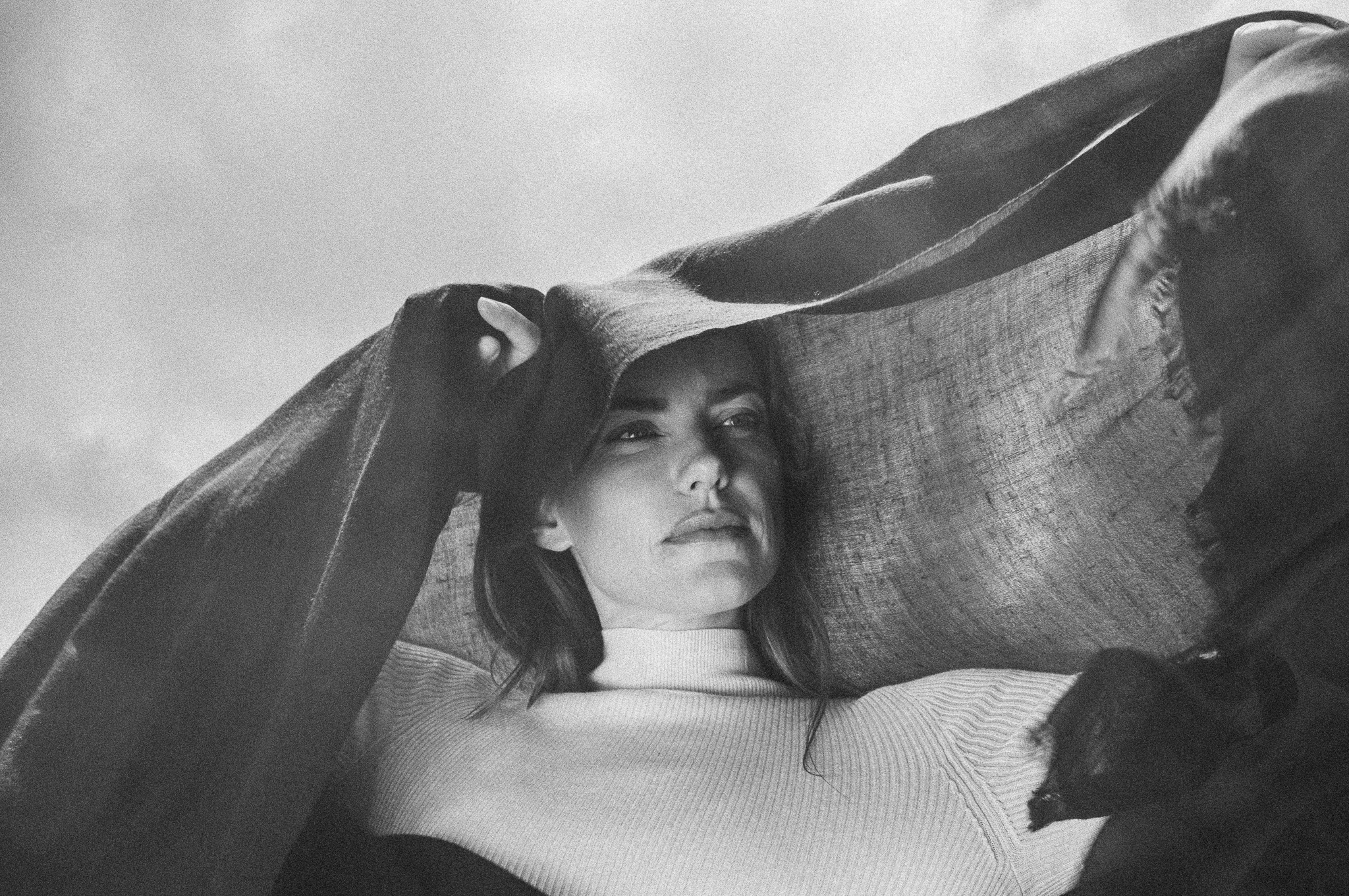 Black and white image of a woman in a white turtleneck holding a pashmina overhead against a cloudy sky. Campaign photography by FADETOBLACK