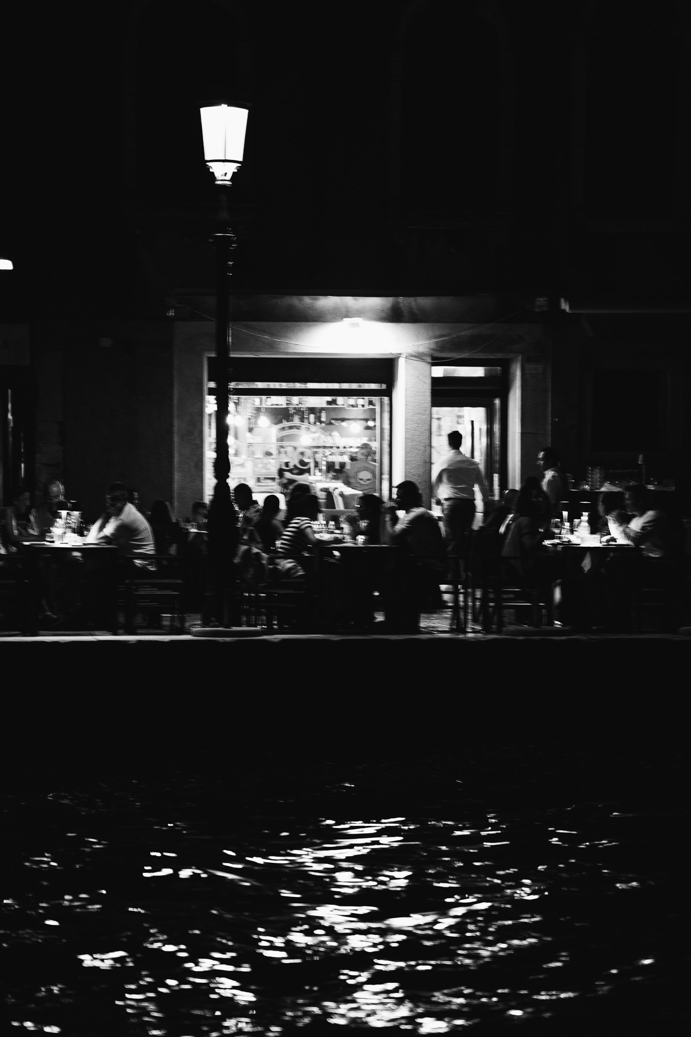 Black and white night scene of a man walking across a dark Venice square past an ornate street lamp. Fine art street photography by FADETOBLACK