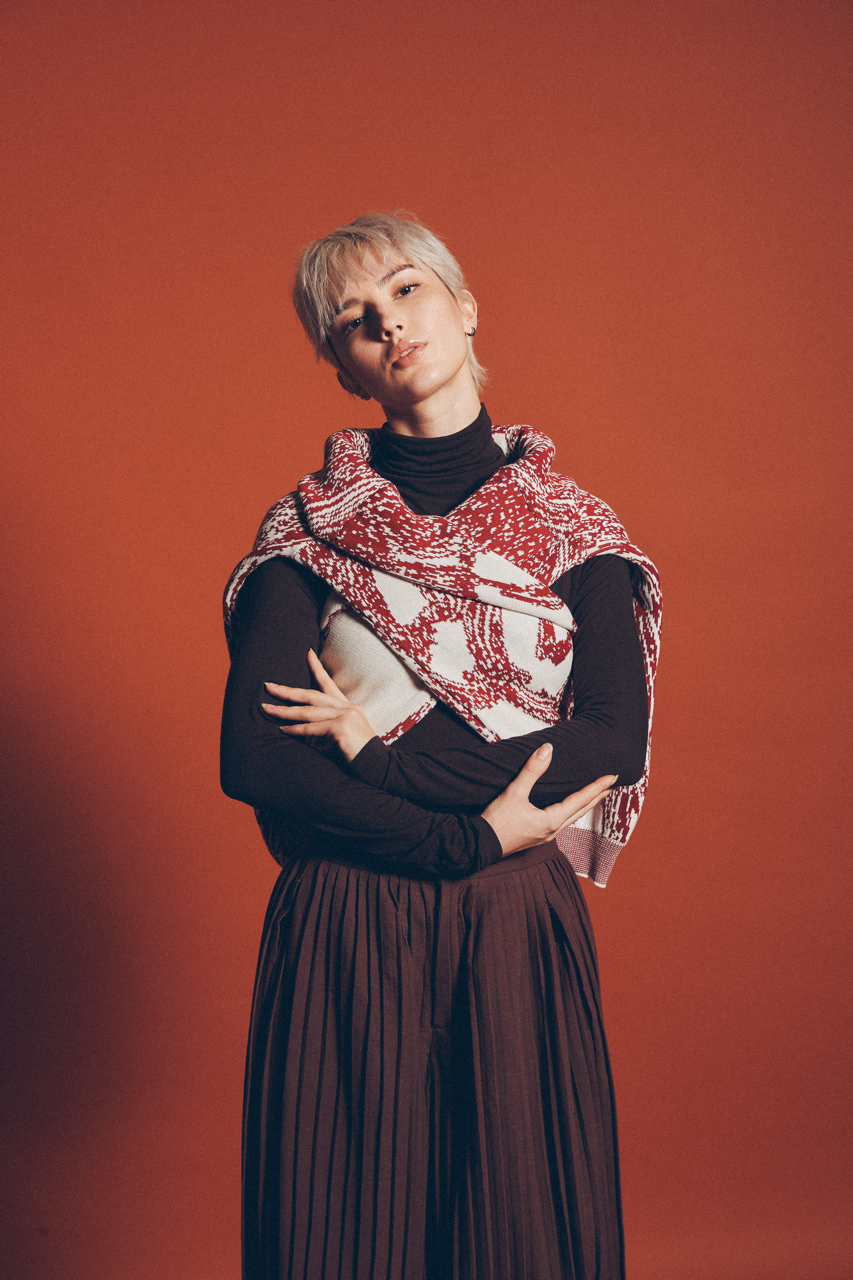 Model with short platinum hair in a red patterned knit wrap and black turtleneck on a rust orange backdrop. Editorial fashion photography by FADETOBLACK