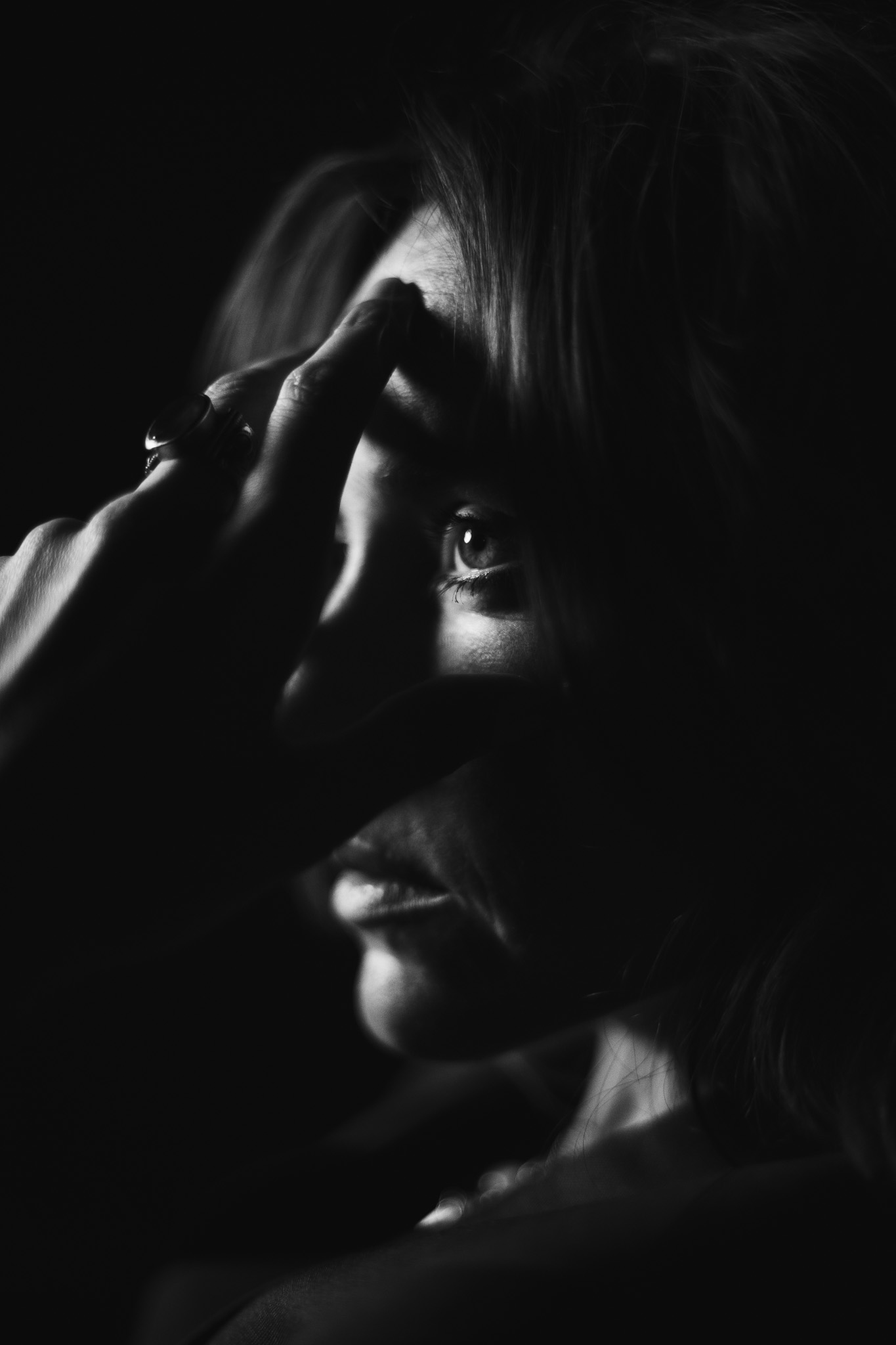 Black and white portrait of a woman hiding her face behind a raised hand with a large ring, deep shadow. Fine art portrait photography by FADETOBLACK