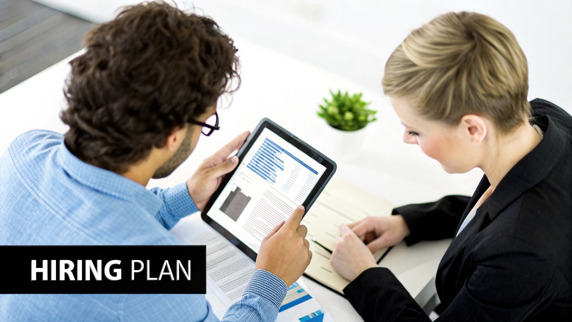 Two colleagues review a hiring plan on a tablet, discussing recruitment strategies in a business meeting.