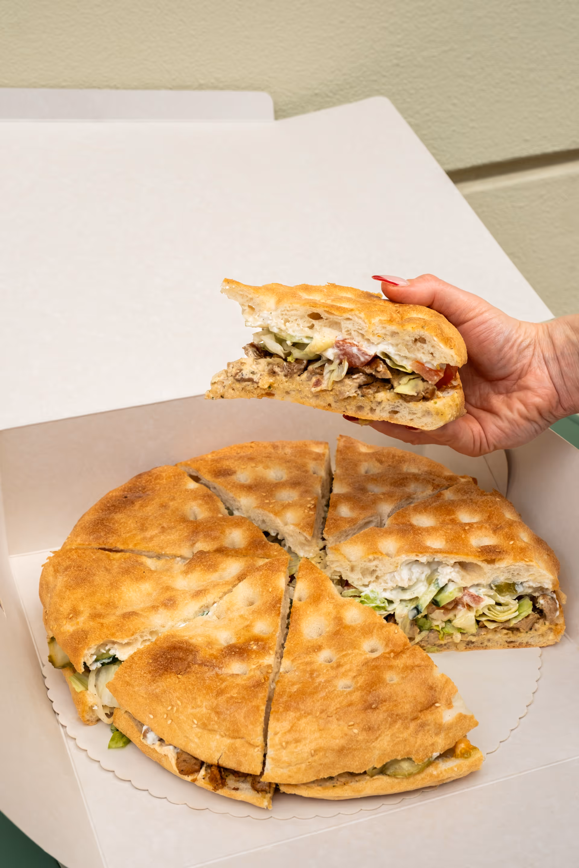 Hand holding a slice of sandwich pie with layers of meat, lettuce, tomato, and sauce inside a round, sliced bread pie.