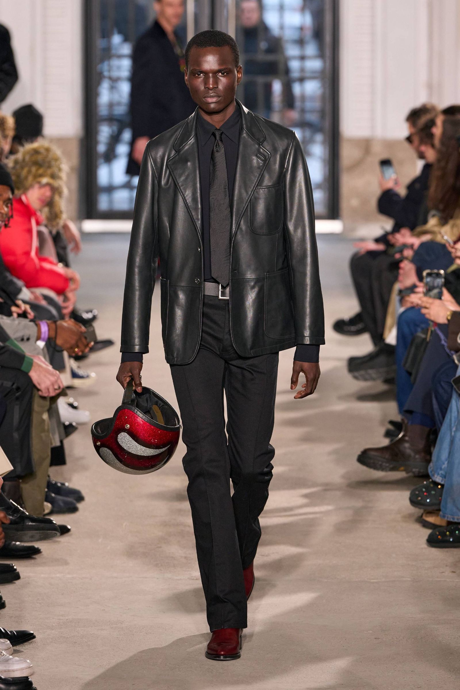 Modelo con casco en mano blazer de piel camisa corbata y pantalón todos en negro con zapatos rojo quemado