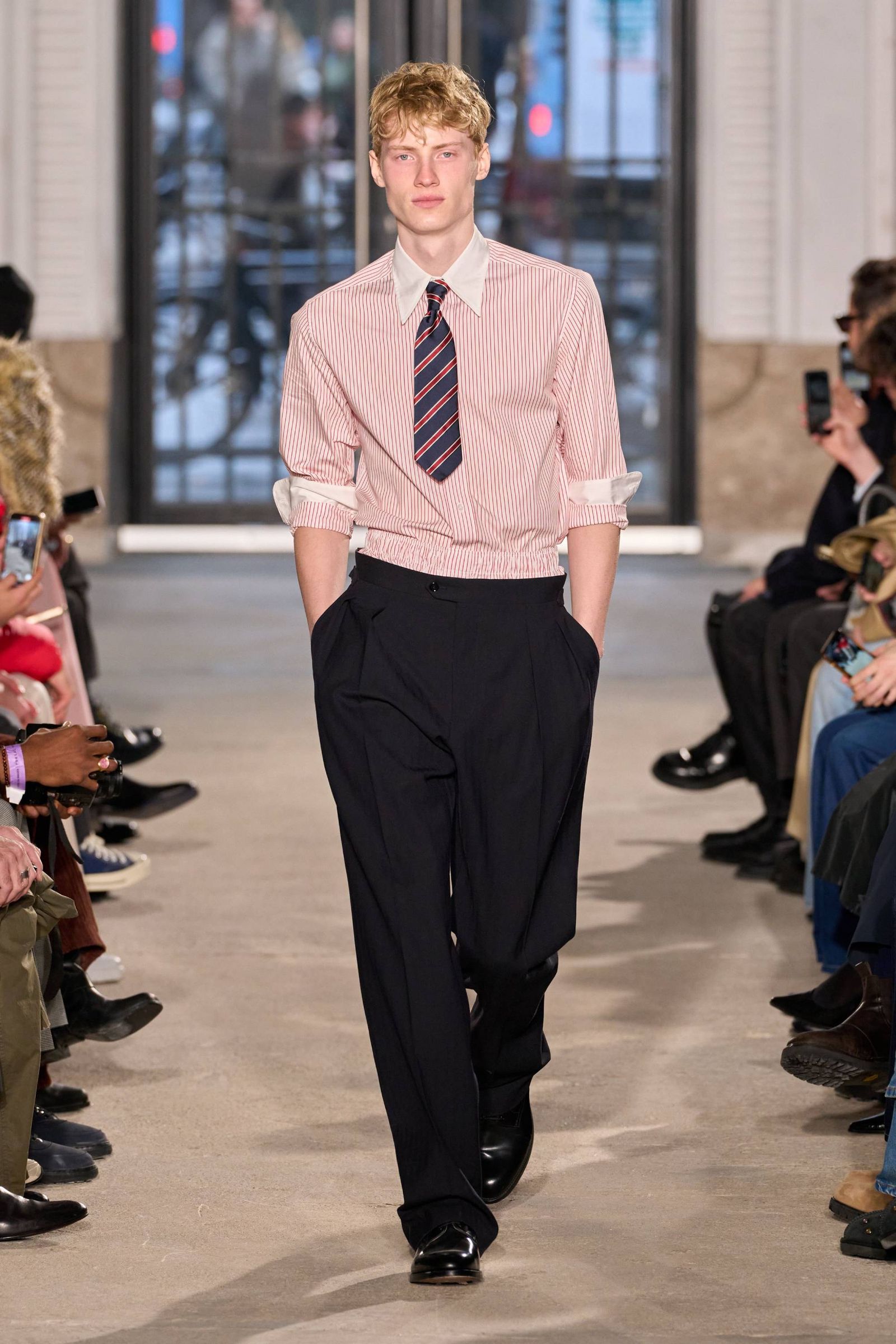 Modelo con camisa rosa a rayas blancas con puños y cuello blanco corbata corta a rayas azul marino y rojo pantalón negro