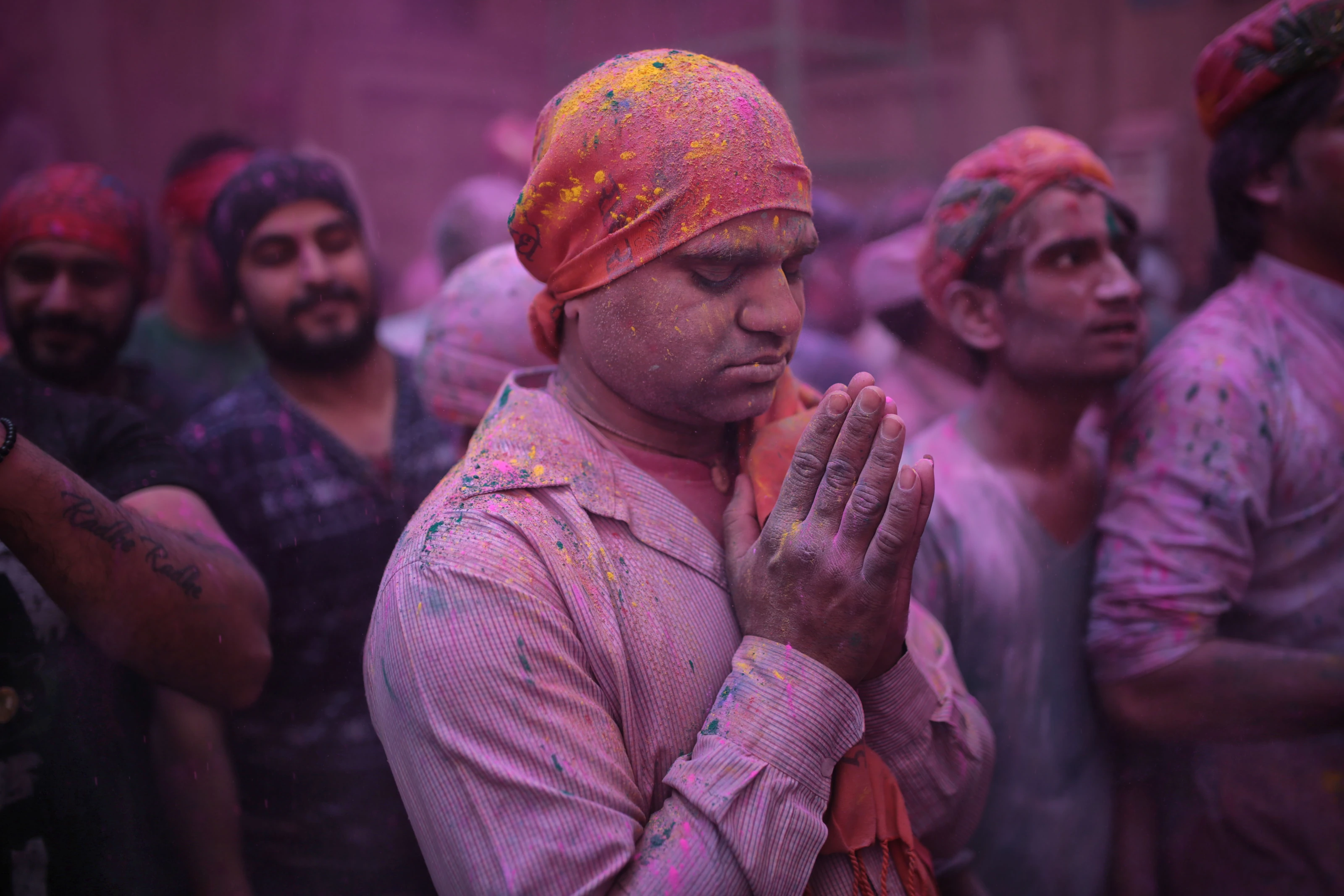 A participant in a pink turban holds his hands together amid a crowd dusted with coloured powder