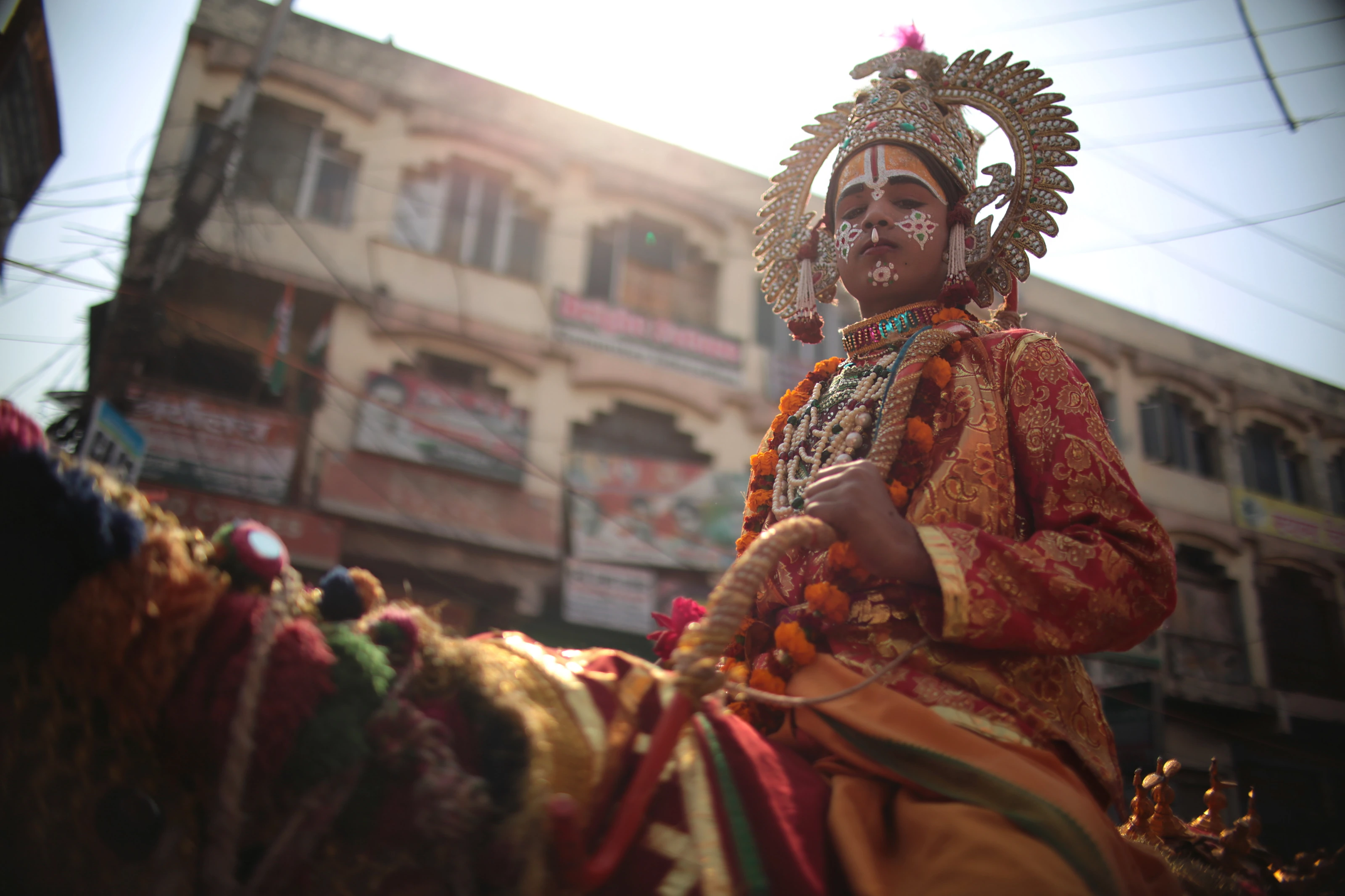A young performer in an ornate red and gold costume rides through a sunlit street procession