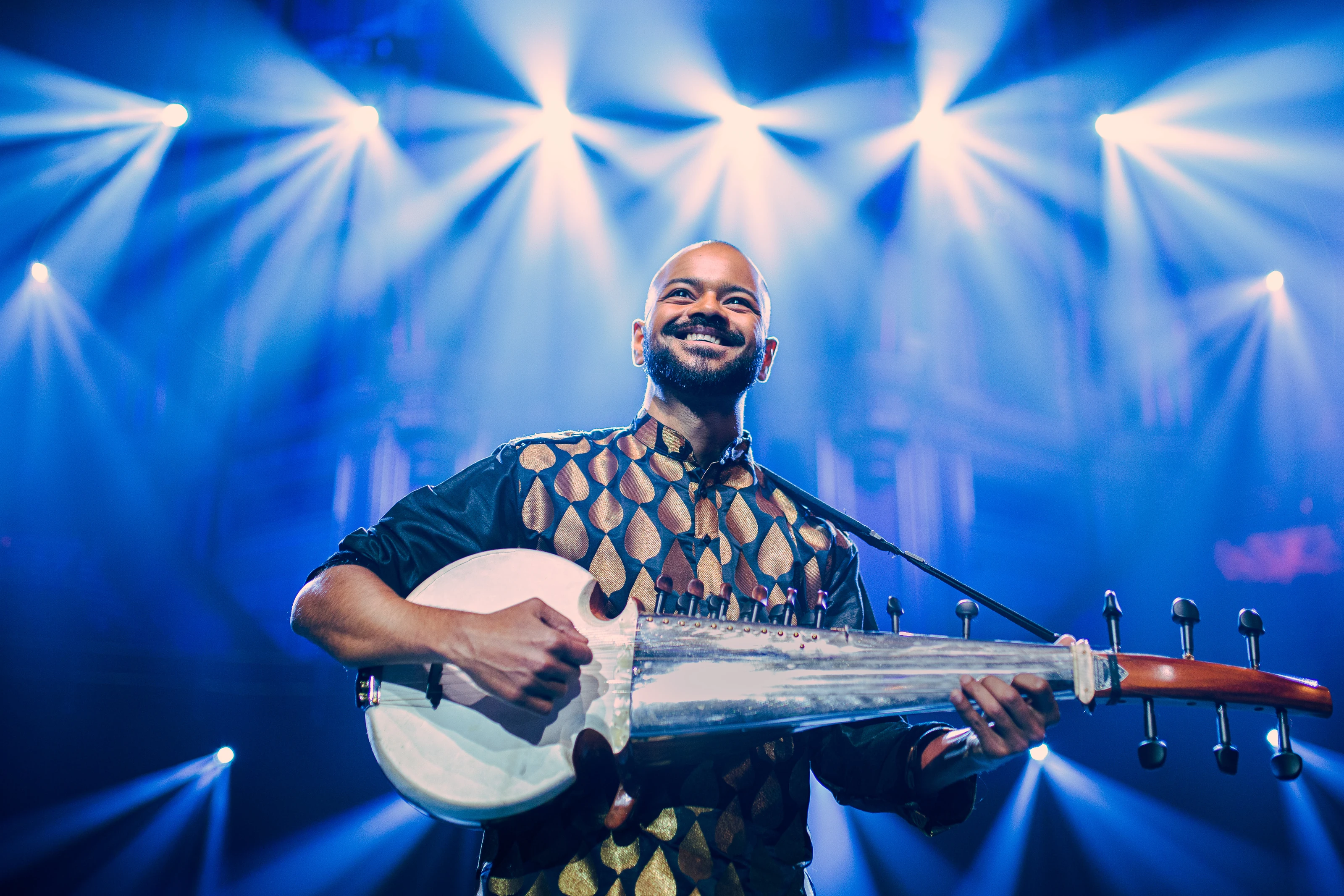 Soumik Datta smiles as he plays the sarod beneath blue stage lights at the Royal Albert Hall.