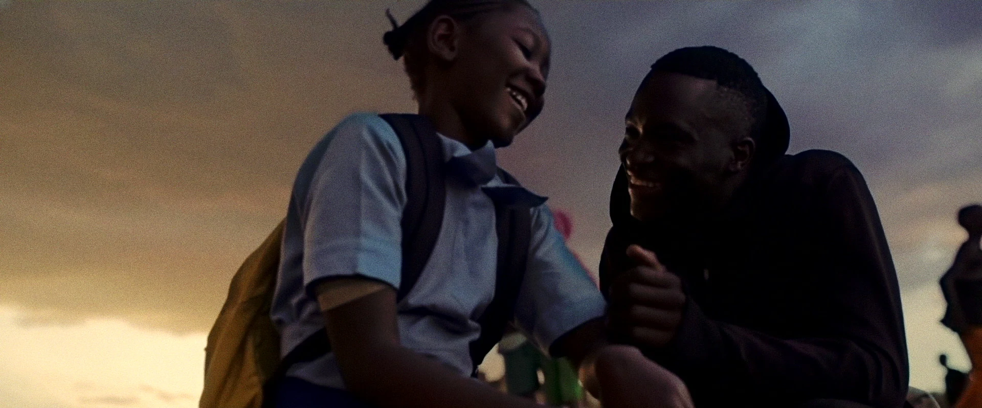 A young girl smiles beside her older brother against an evening sky