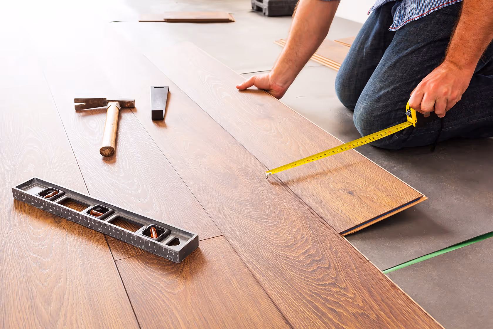 Person kneeling on floor measuring wooden flooring plank with a tape measure, with hammer and level nearby.