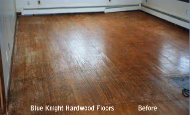 Worn hardwood floor in an empty corner room before refinishing.
