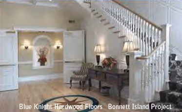 Spacious living room with wooden floors, patterned armchairs, side table with lamps, floral decorations, and white staircase with railing.