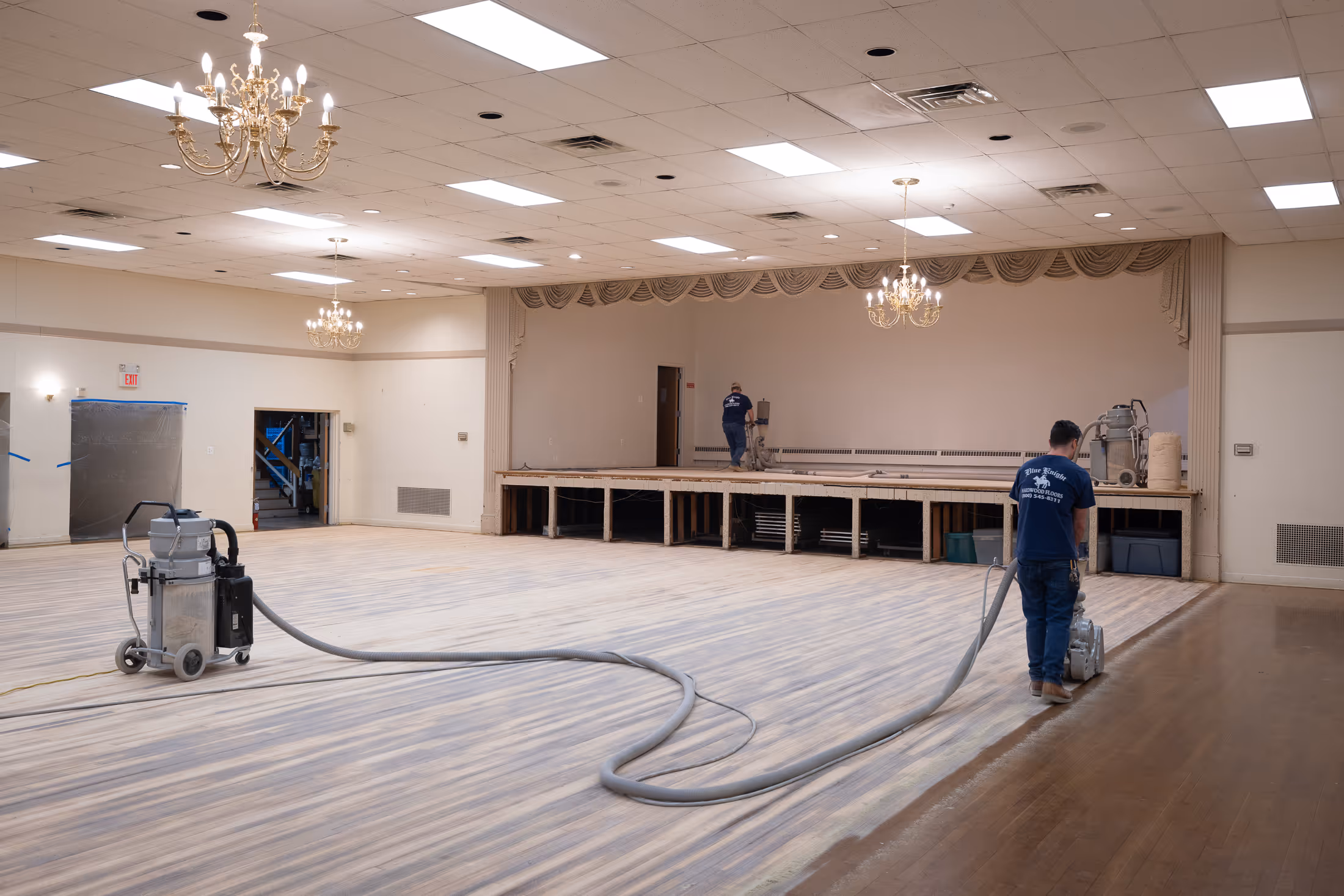 Two workers refinishing a large wooden ballroom floor with sanding machines under chandeliers.