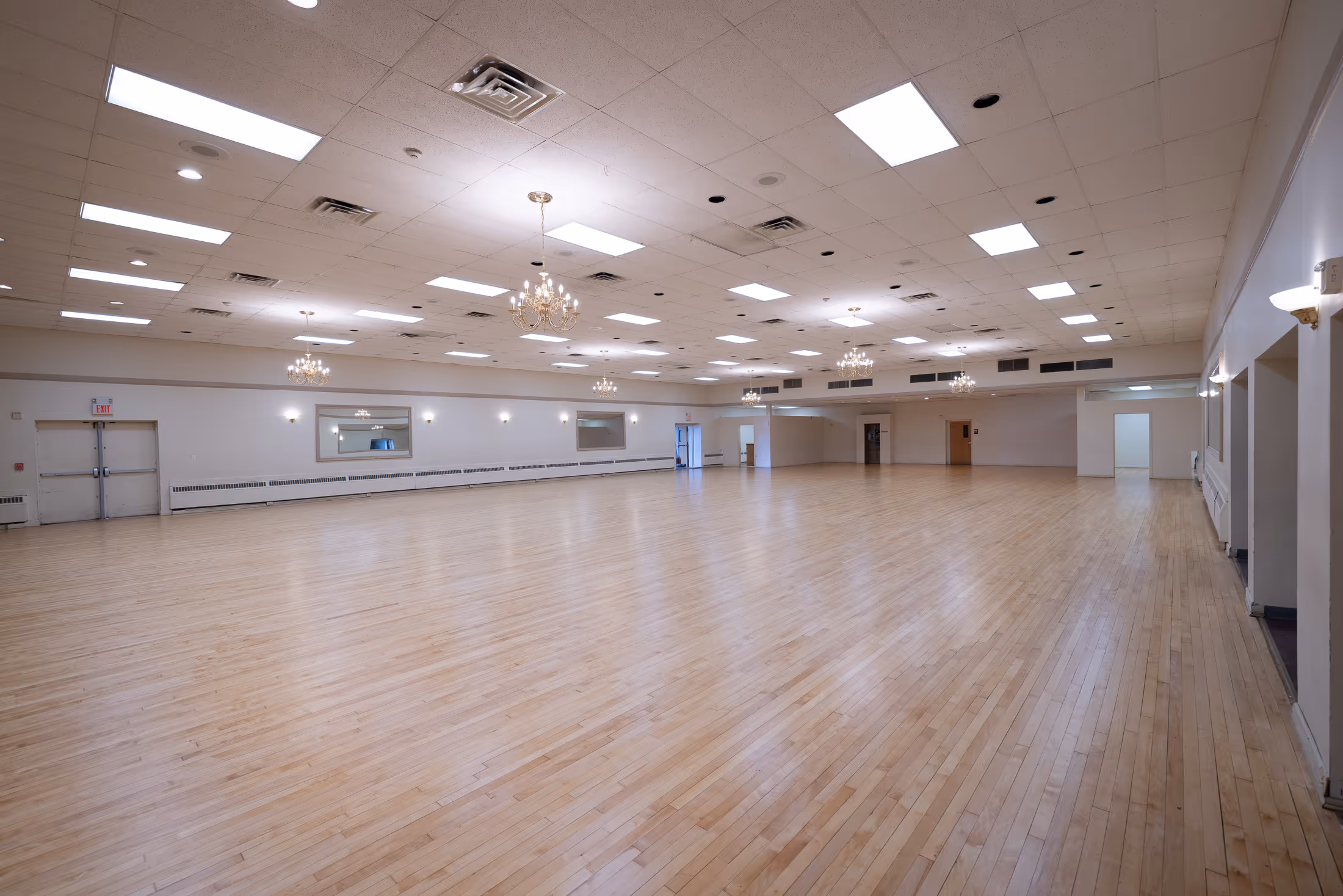 Large empty ballroom with polished wooden floor, white walls, chandeliers, and ceiling lights.
