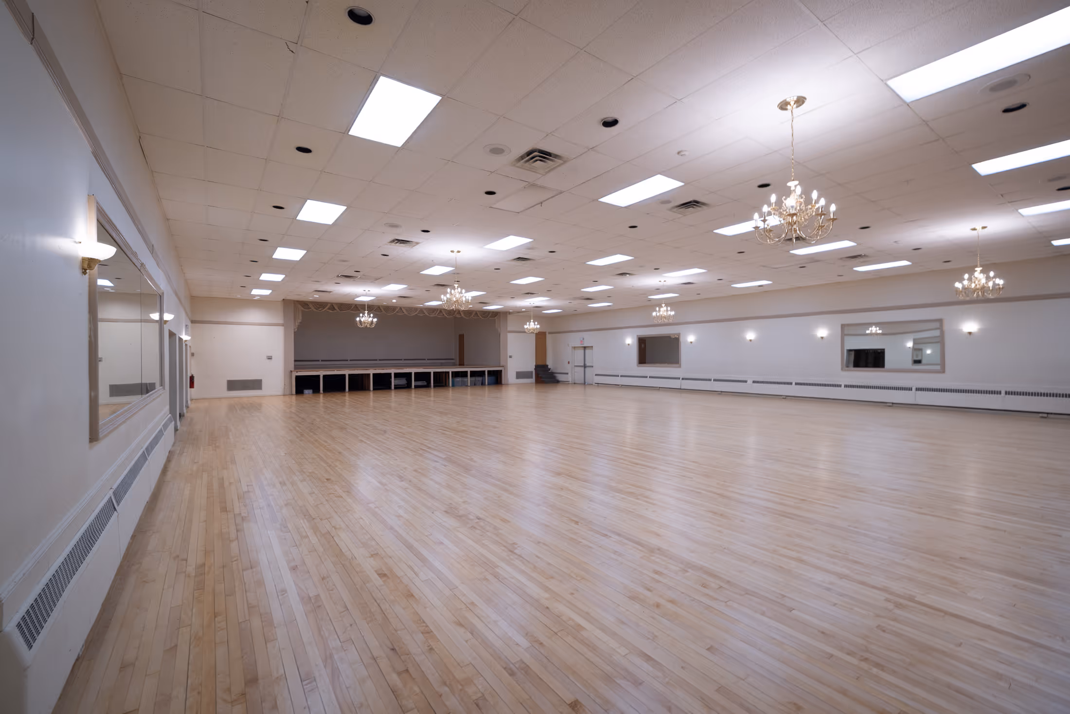 Large empty ballroom with polished light wood floor, chandeliers, and wall mirrors under a white ceiling with recessed lighting.