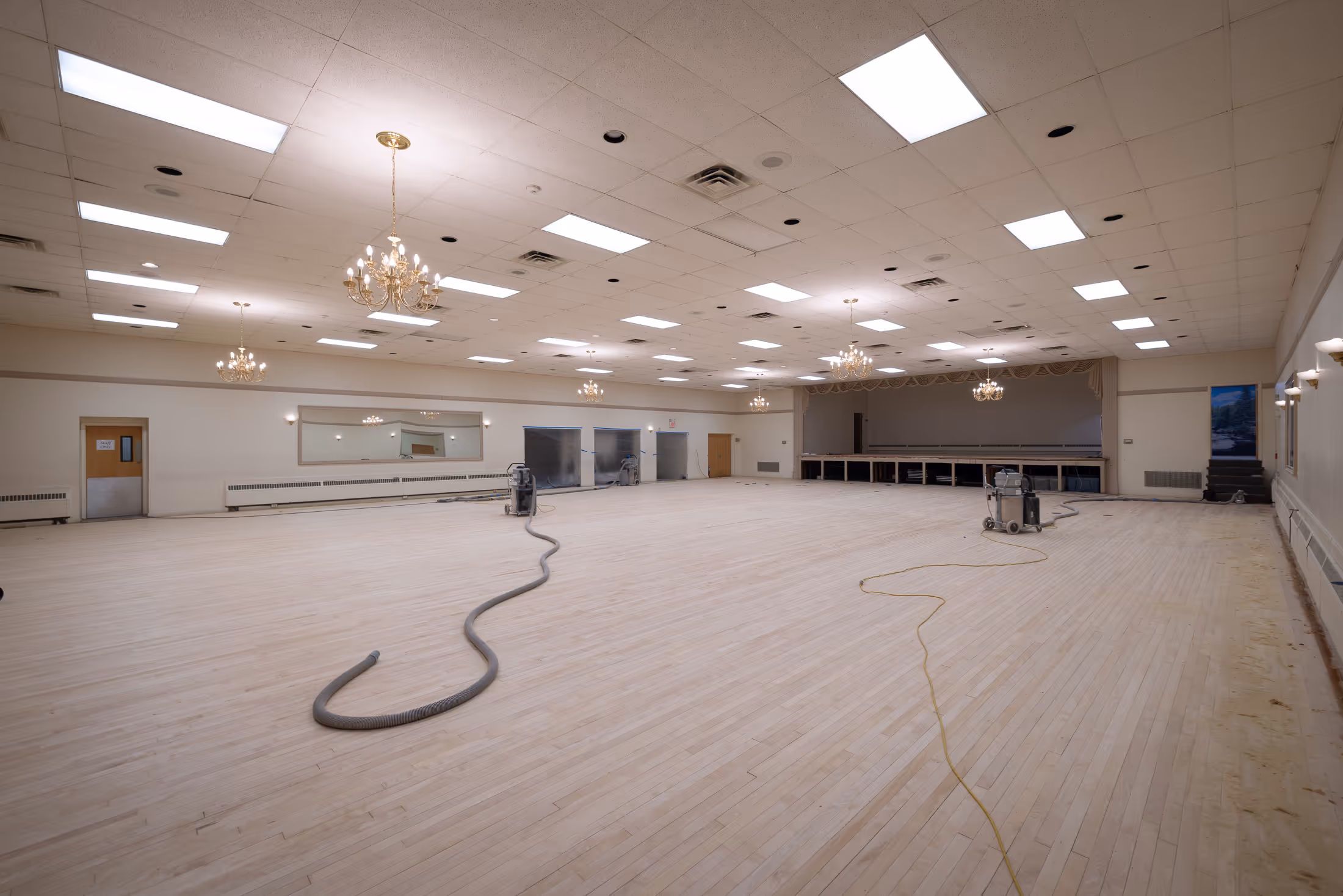 Large empty ballroom with light wooden floor being refinished, chandeliers hanging from ceiling, and floor sanding equipment in the room.