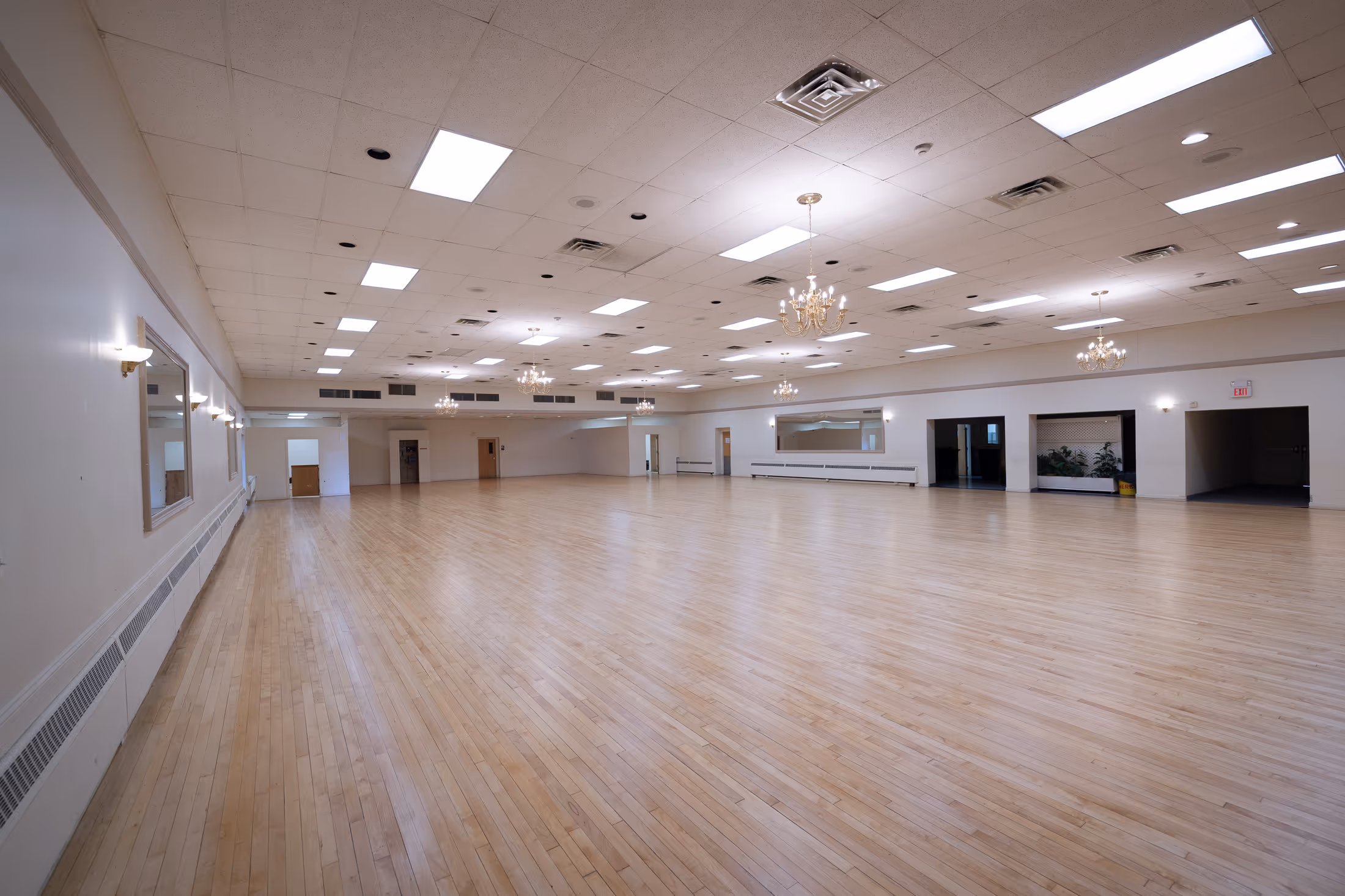 Spacious empty ballroom with polished hardwood floor, white walls, chandeliers, and ceiling lights.