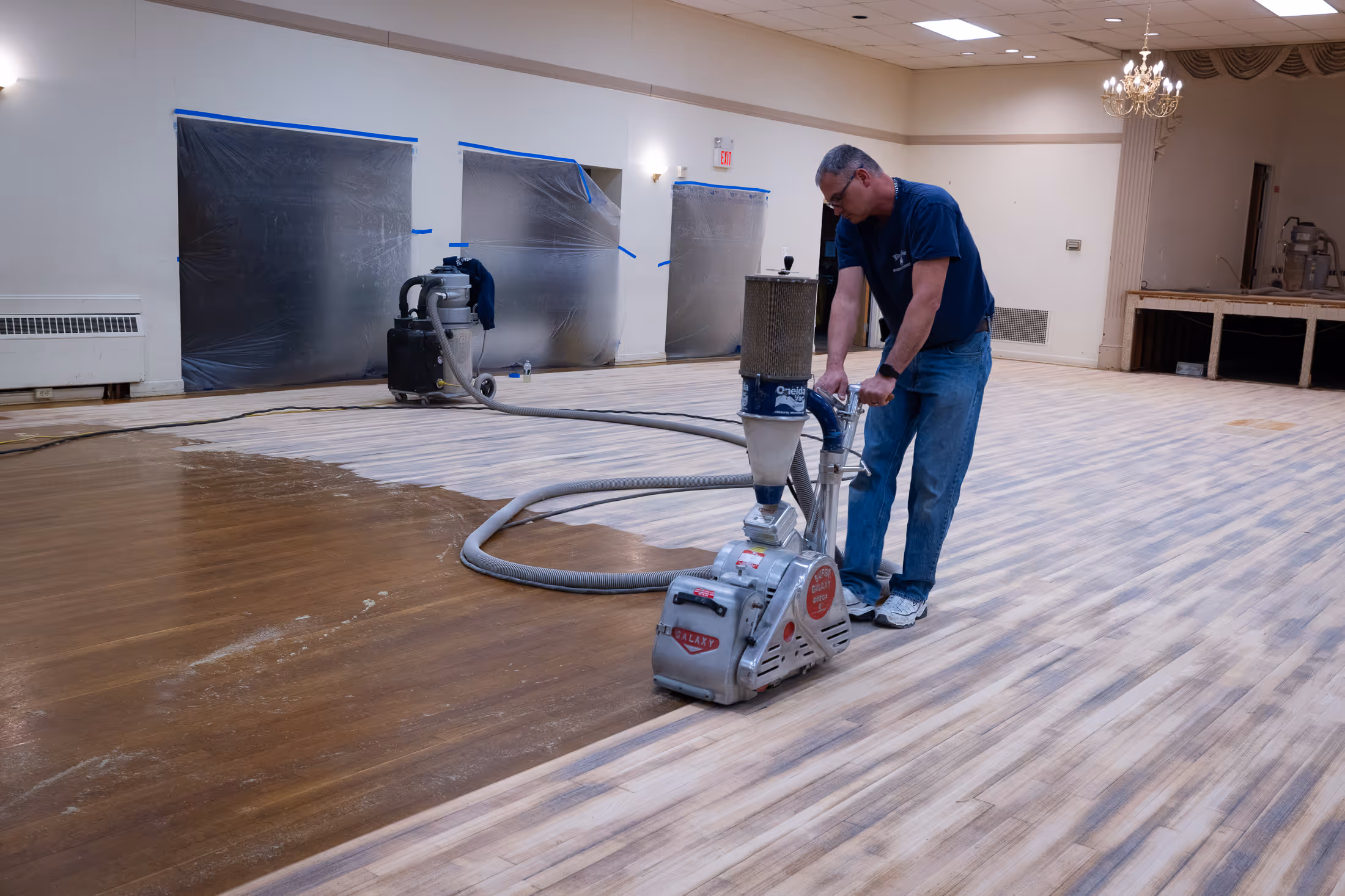 Man refinishing a wooden floor using a sanding machine in a large room with plastic-covered windows.