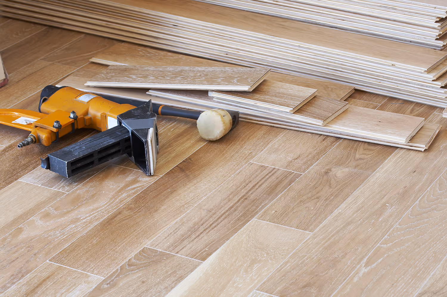 Stack of uninstalled wooden flooring planks with a pneumatic flooring nailer and rubber mallet on a partially installed hardwood floor.