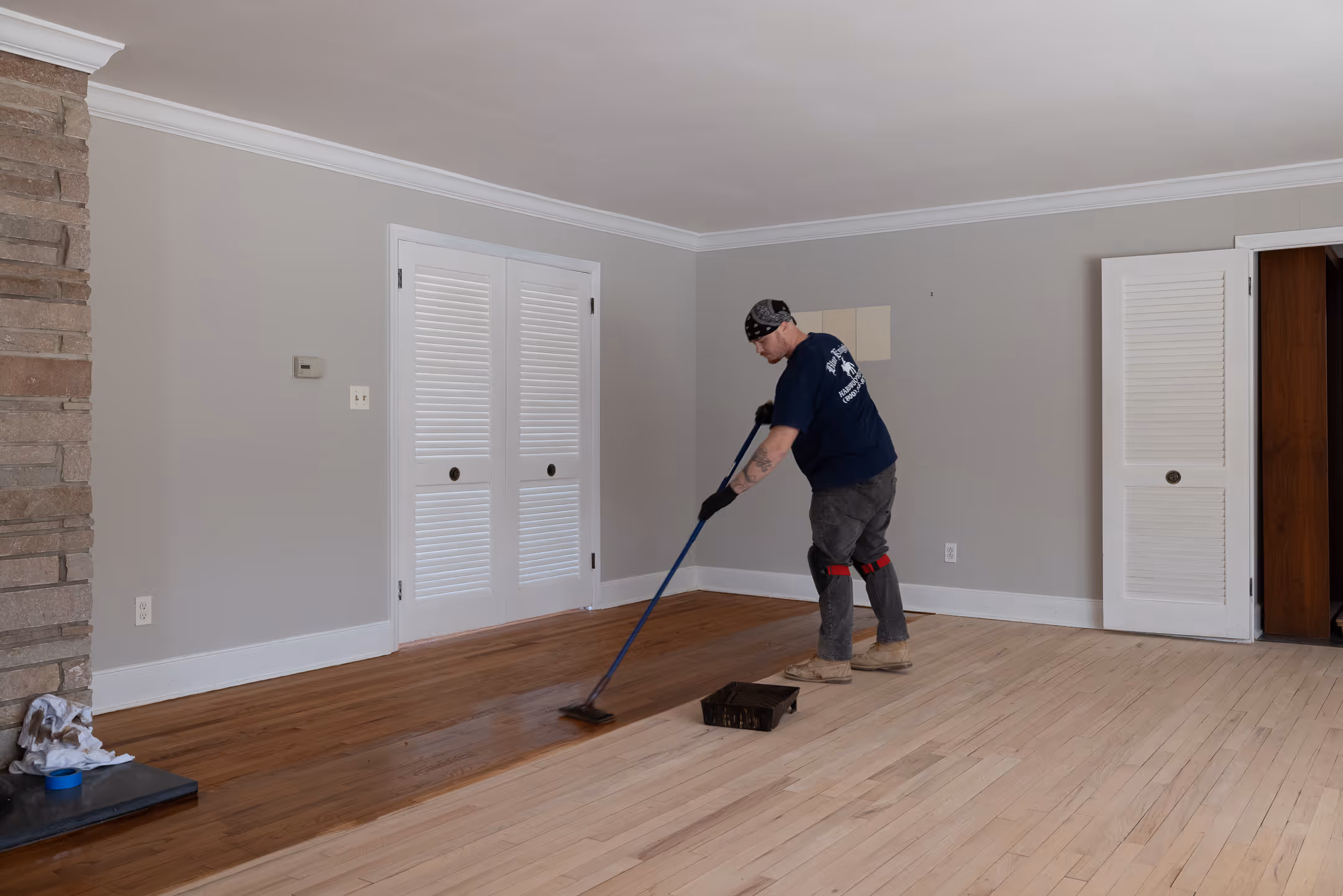 Man applying wood stain to a hardwood floor in a room with gray walls and white doors.