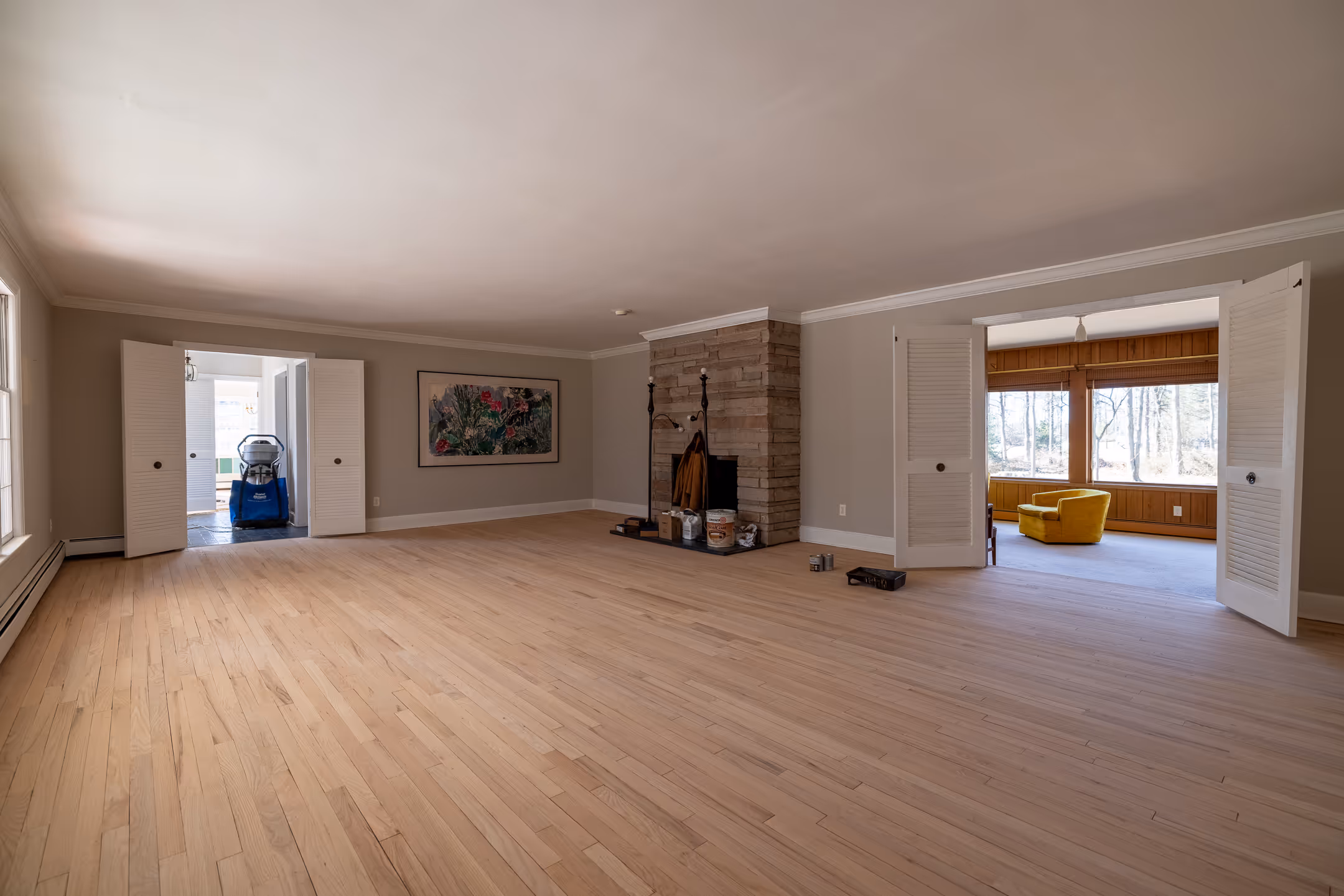 Spacious room with light hardwood floors, stone fireplace, and open white louvered doors leading to adjacent rooms, one with a yellow armchair.