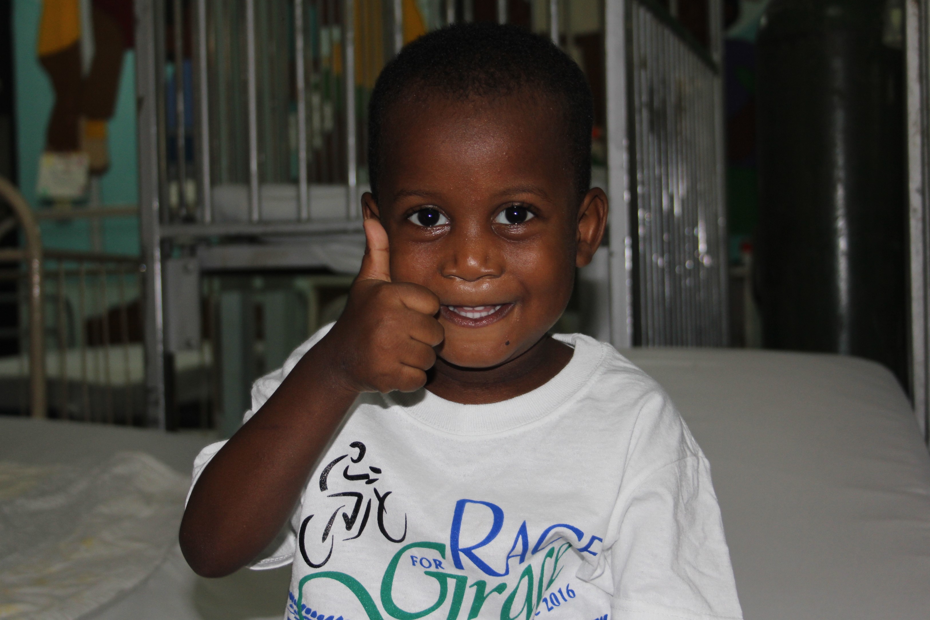 Smiling young child in a hospital bed giving a thumbs-up gesture.