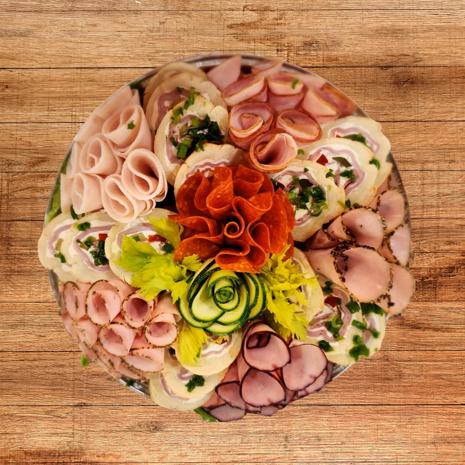 Round platter with assorted sliced deli meats, pinwheel sandwiches, and garnished with celery and cucumber slices on a wooden surface.
