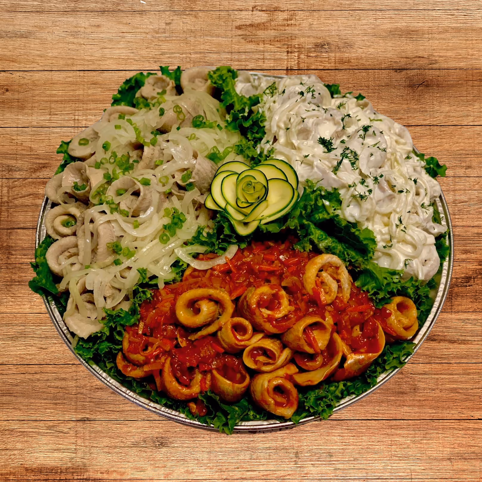 Round platter with three sections of food: sliced meat with onions and scallions, creamy salad with dill, and rolled pieces in red sauce, all on a bed of lettuce with a cucumber rose in the center.
