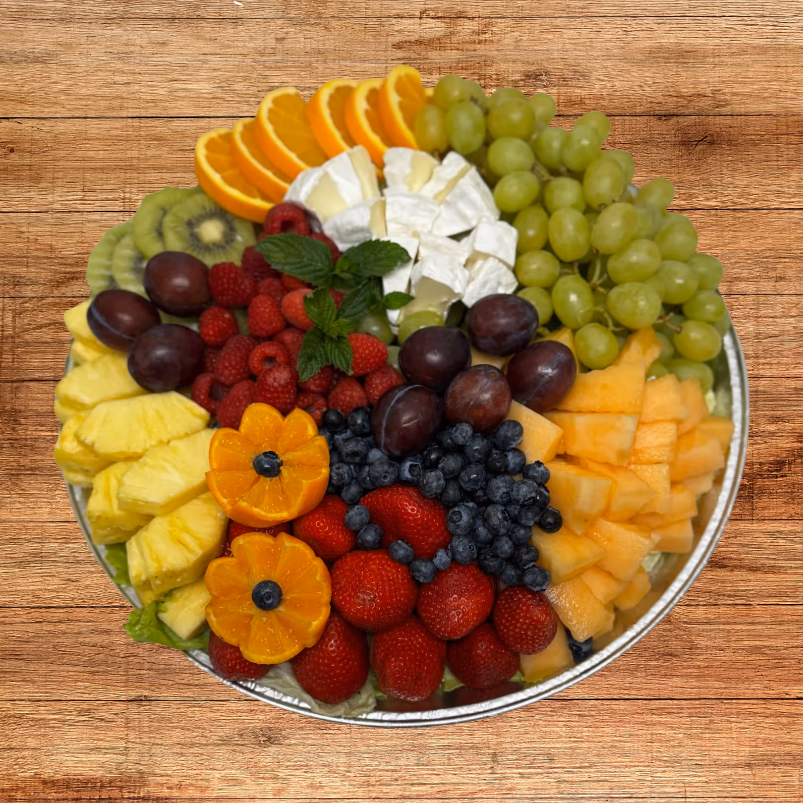 Round platter with a colorful assortment of sliced oranges, green grapes, cantaloupe, blueberries, strawberries, raspberries, plums, pineapple, kiwi, two halved kumquats with blueberries, and wedges of Brie cheese garnished with mint leaves.