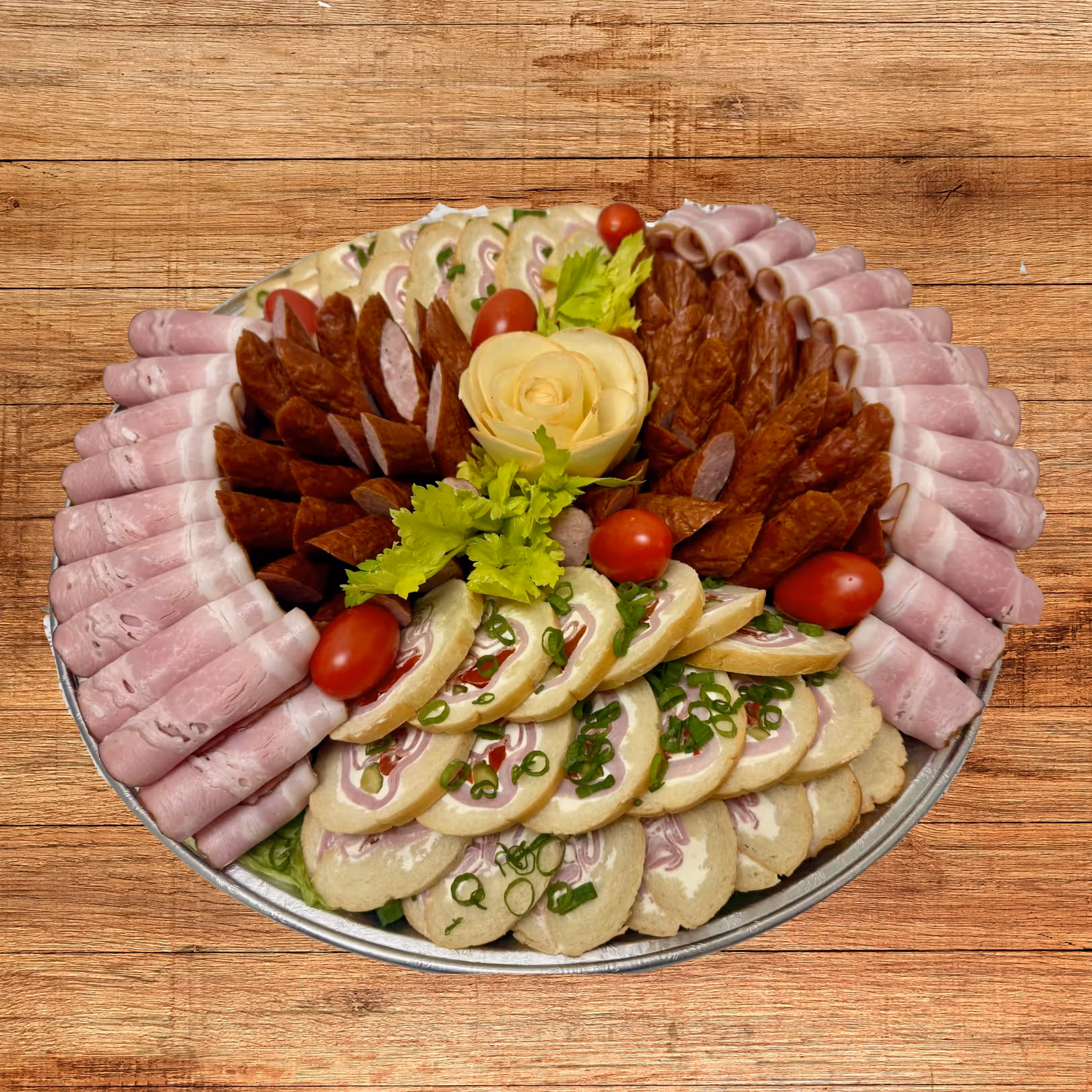 Round platter with assorted deli meats including rolled ham, sliced sausage, and cream cheese pinwheels garnished with cherry tomatoes and celery leaves.
