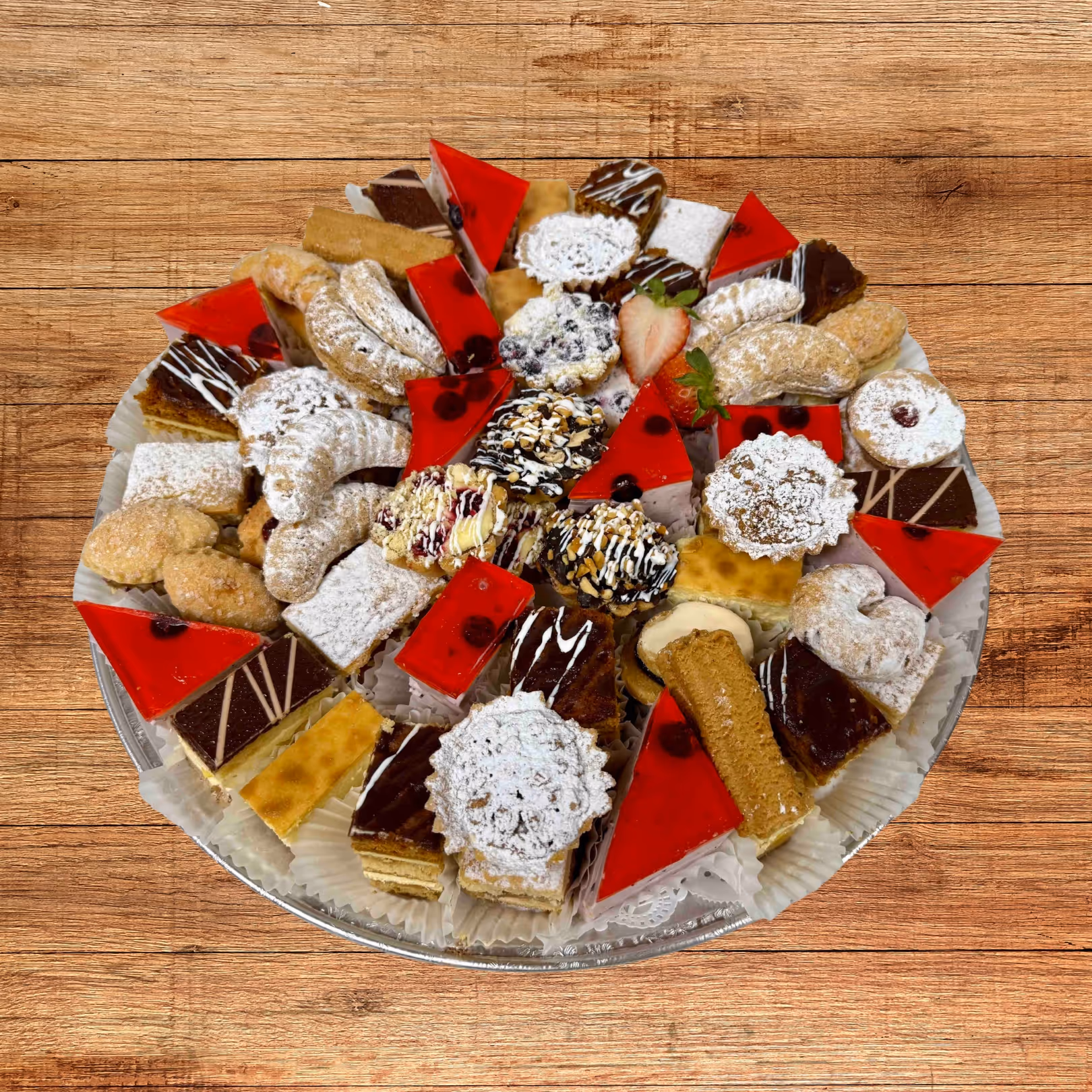 Assorted plate of various colorful European-style pastries and cookies, including powdered sugar crescents, red jelly-topped cakes, and nut-covered treats on a white paper doily.