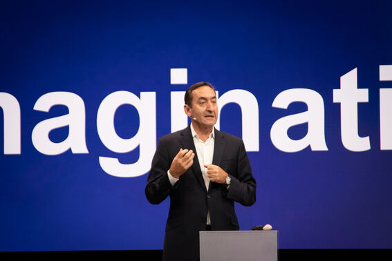 Person in a dark suit giving a presentation in front of a large screen displaying the word 'imagination'.