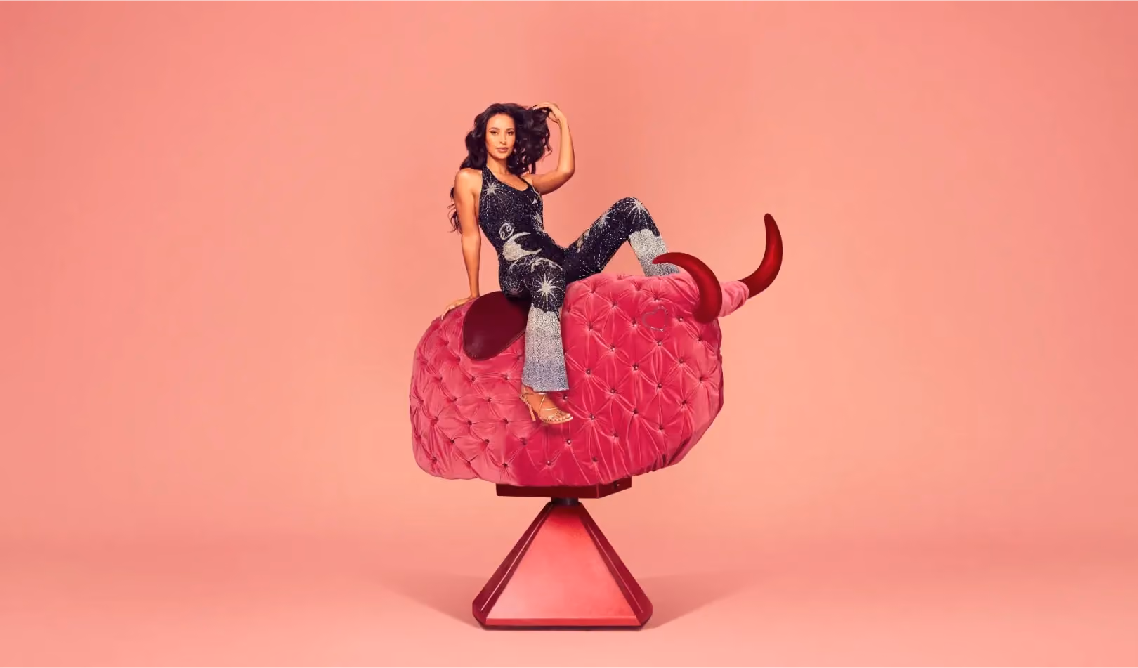 Love Island host Maya Jama lounges on the back of a hot pink velour mechanical bull. She flicks her hair back, looking at the camera looking confident and glamorous.
