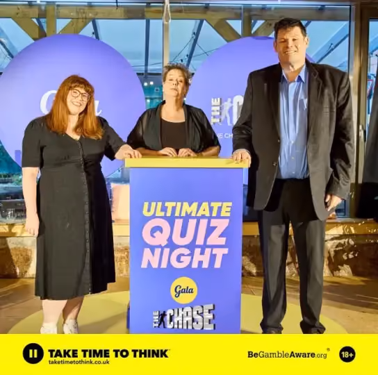 Three people stand behind a podium with 'Ultimate Quiz Night Gala The Chase' sign in a quiz event setting.