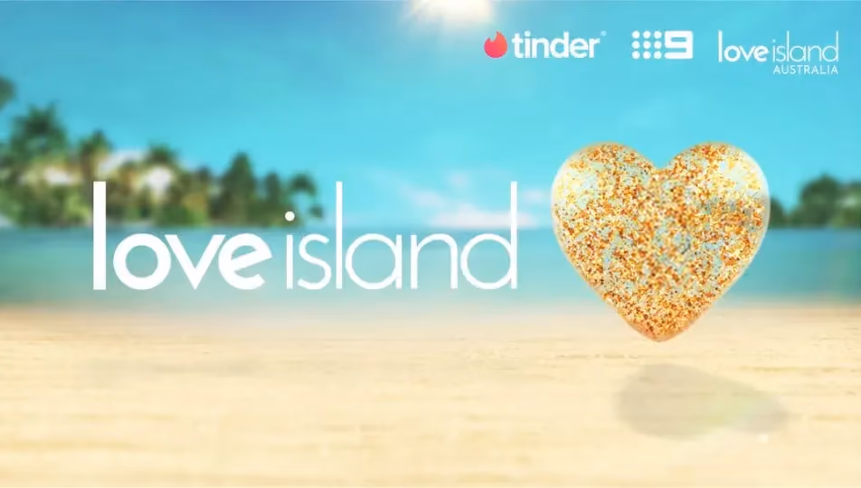Love Island logo with a golden heart on a sandy beach under bright blue sky and sun.