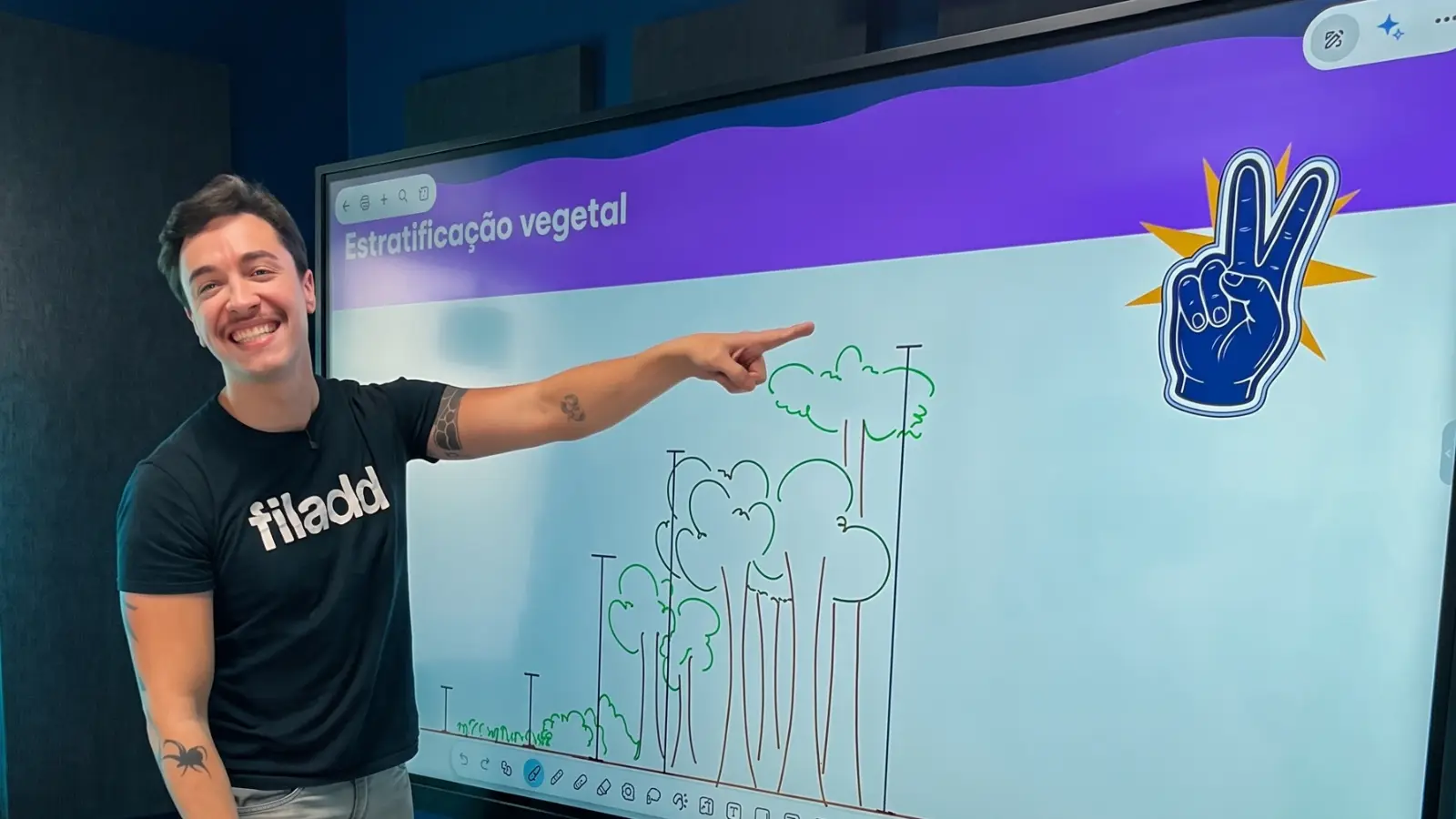 Prof. Rafa gravando aula de Biologia no estúdio da Filadd, apontando para um esquema de estratificação vegetal na lousa digital