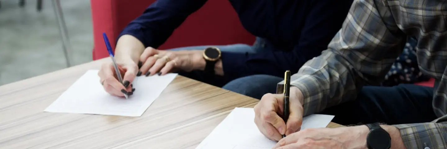 Two people sitting at a table signing documents, with glasses and a wallet placed nearby.