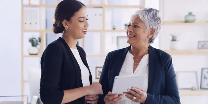 Two businesswomen in an office discussing information on a digital tablet.
