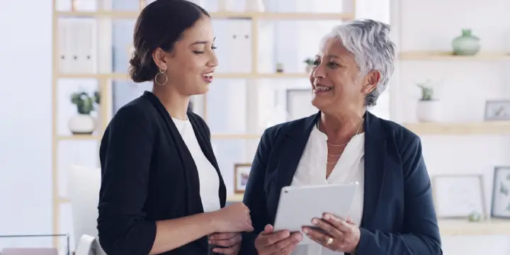 Two businesswomen in an office discussing information on a digital tablet.