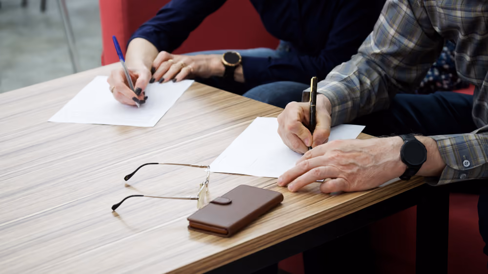 Two people sitting at a table signing documents, with glasses and a wallet placed nearby.
