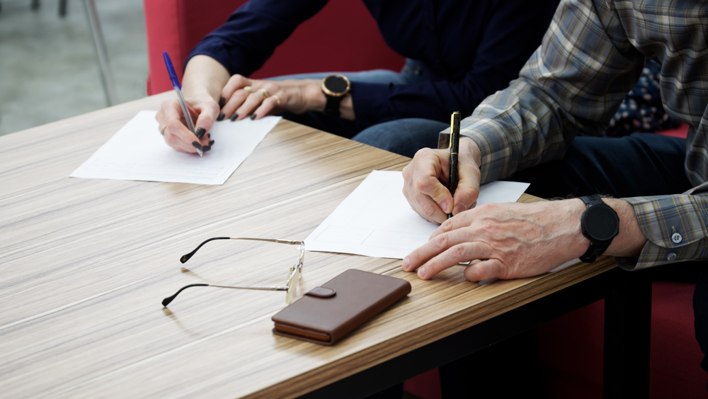 Two people sitting at a table signing documents, with glasses and a wallet placed nearby.