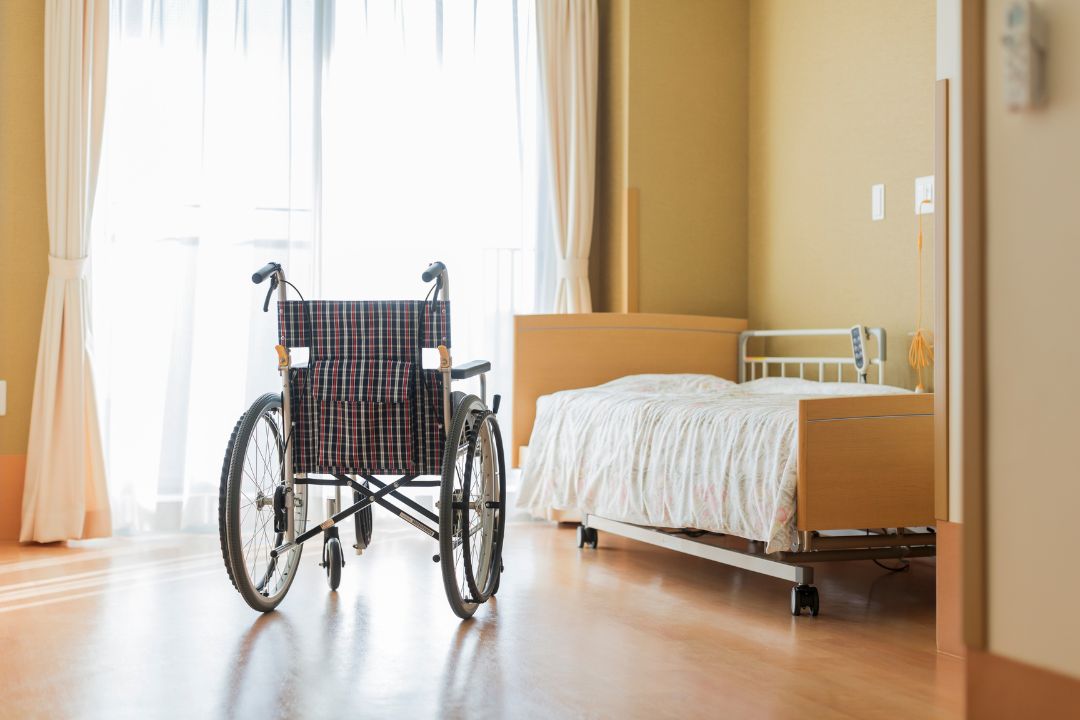 Empty wheelchair facing a hospital bed in a bright patient room with wooden flooring and white curtains.