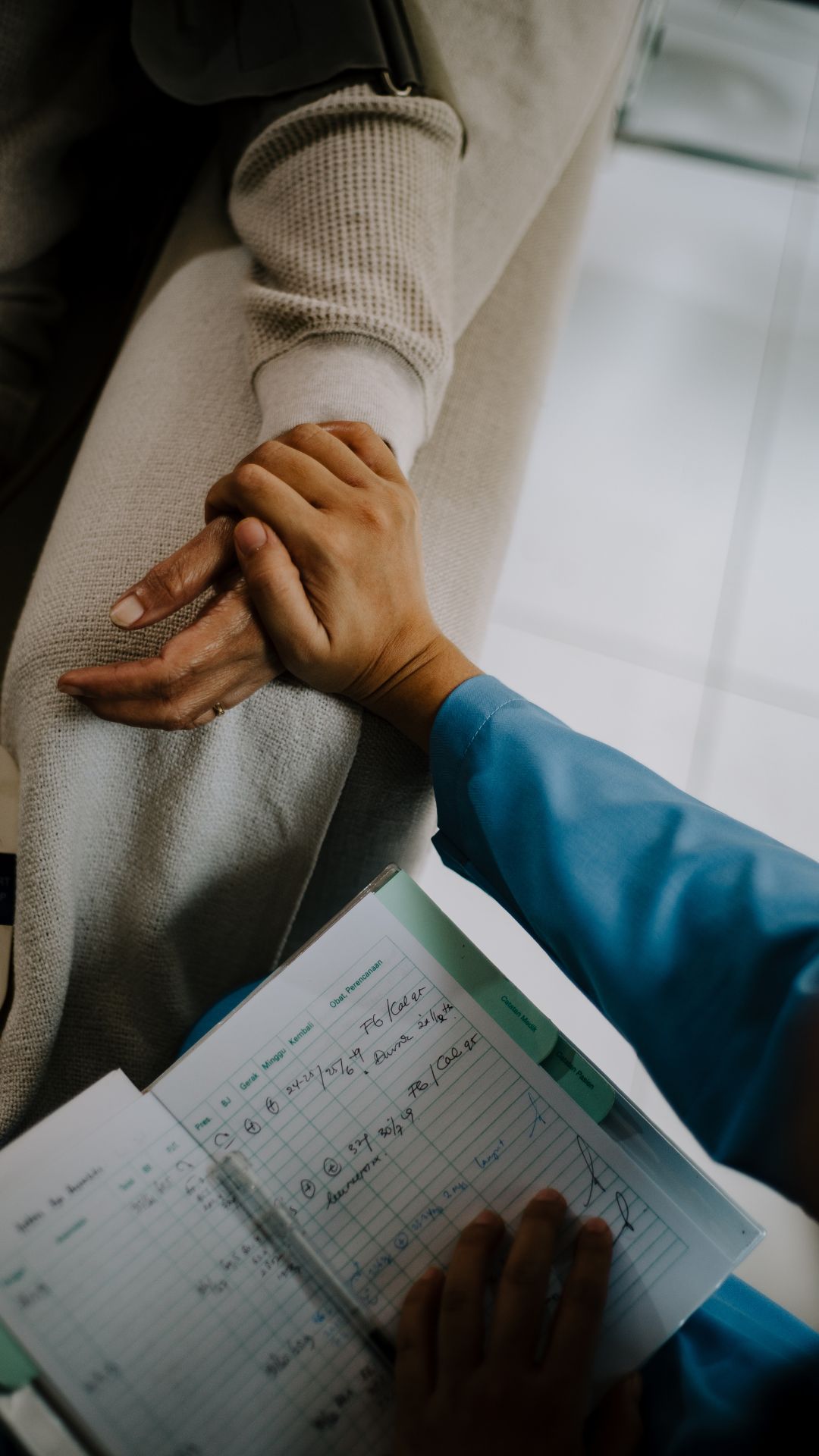 A person in blue scrubs holding a patient's hand while reviewing medical notes on a clipboard.