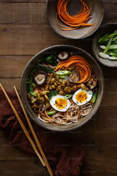 Bowl of ramen noodles topped with boiled eggs, shiitake mushrooms, chopped scallions, crispy bits, and carrot ribbons, with chopsticks beside the bowl on a wooden table.