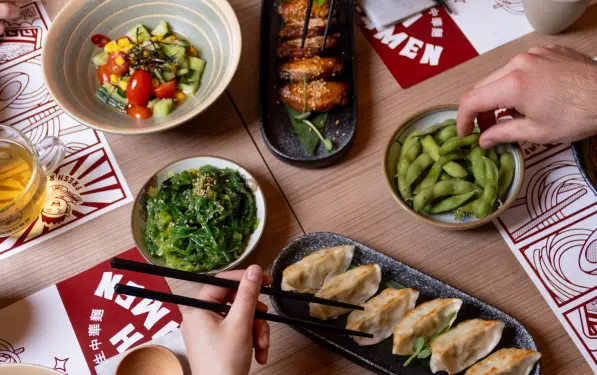 Table with various Japanese dishes including dumplings, edamame, seaweed salad, cucumber and tomato salad, grilled chicken skewers, and a cup of tea with hands holding chopsticks and picking edamame.