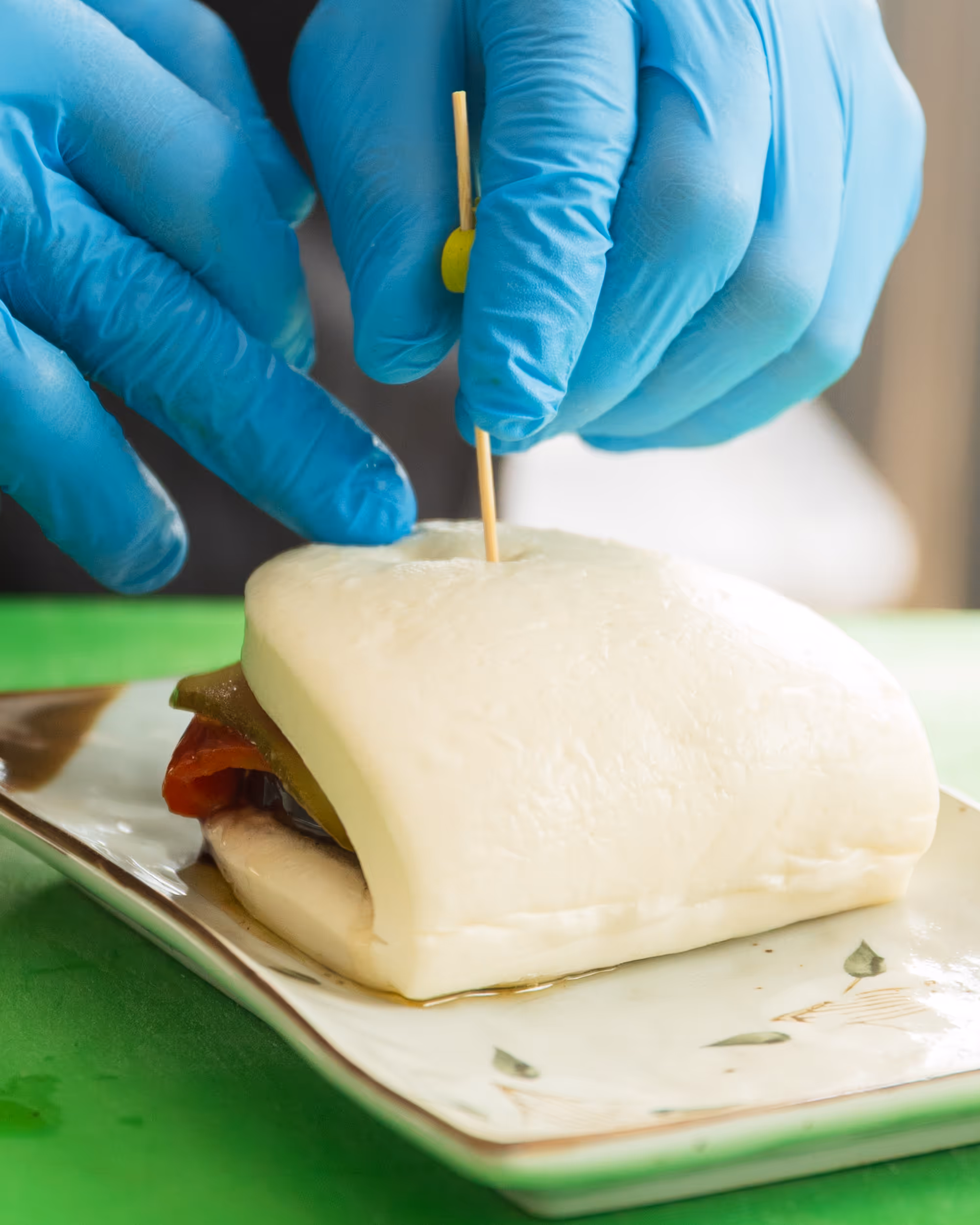 Hands wearing blue gloves securing a skewer into a sandwich with soft white bread and visible tomato and pickles on a plate.