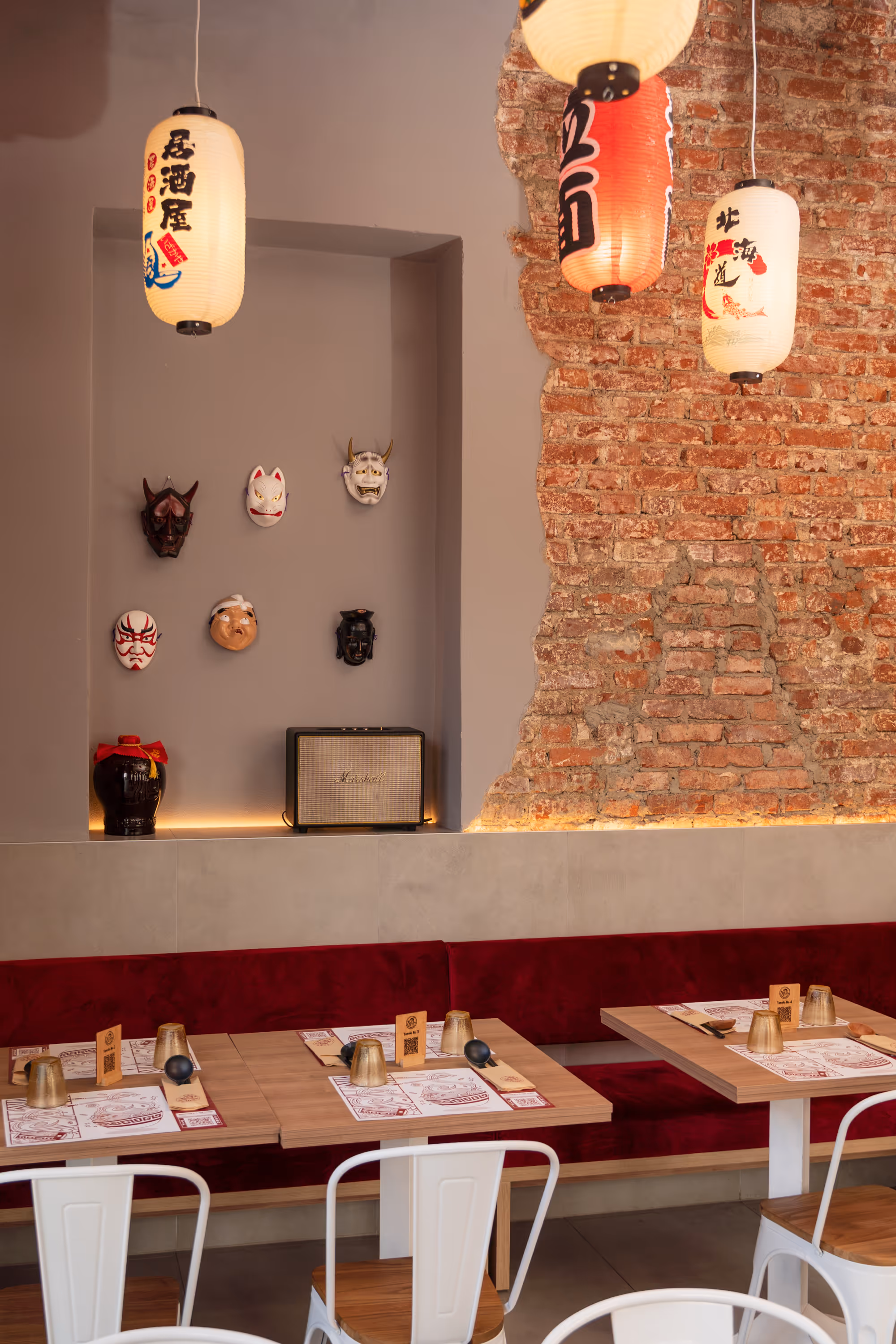 Cozy restaurant interior with wooden tables, white chairs, red cushioned bench, hanging Japanese lanterns, and decorative Japanese masks on a gray wall next to a brick wall.