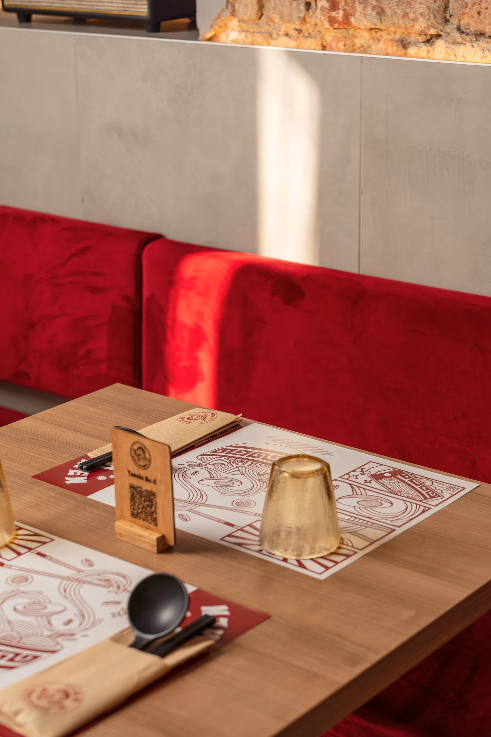 Wooden restaurant table set with paper placemats, wrapped cutlery, a black spoon, an upside-down glass, and a wooden table number holder against a red cushioned bench.