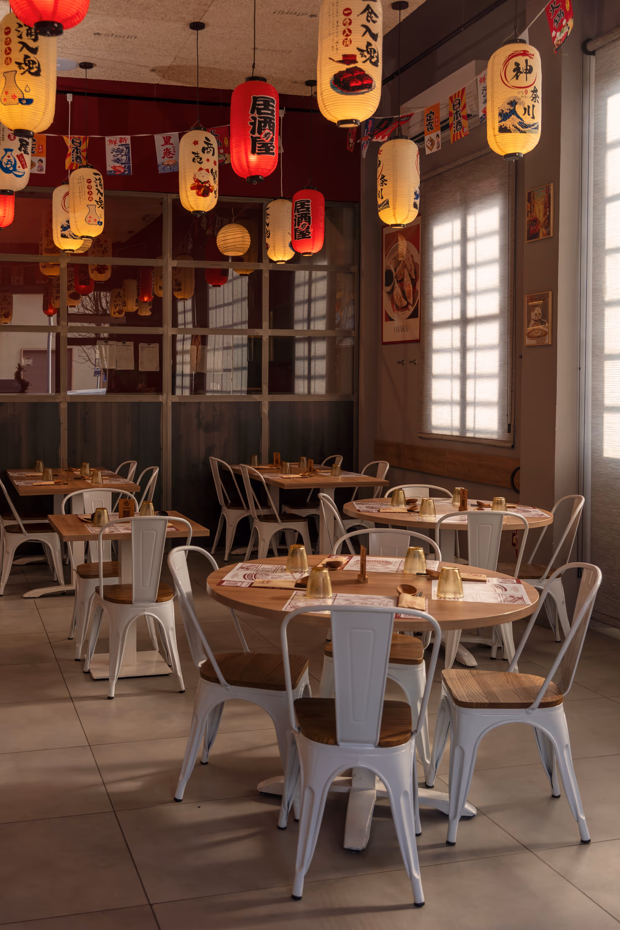 Cozy Japanese restaurant interior with wooden tables, white metal chairs, and hanging red and white paper lanterns featuring Japanese characters.