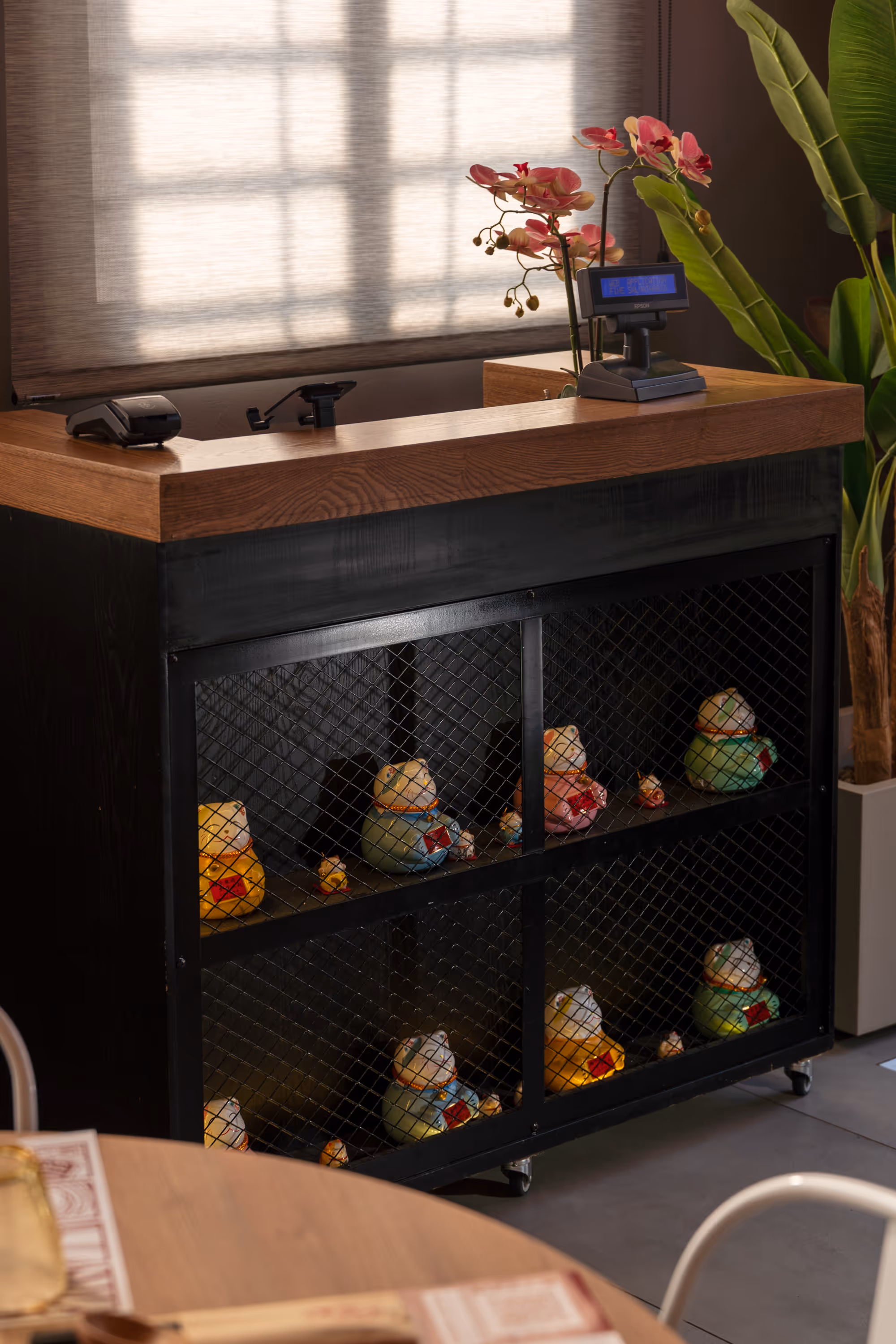 Reception desk with wooden countertop, pink orchid plants, and black metal shelf below displaying colorful ceramic cat figurines behind a wire mesh.